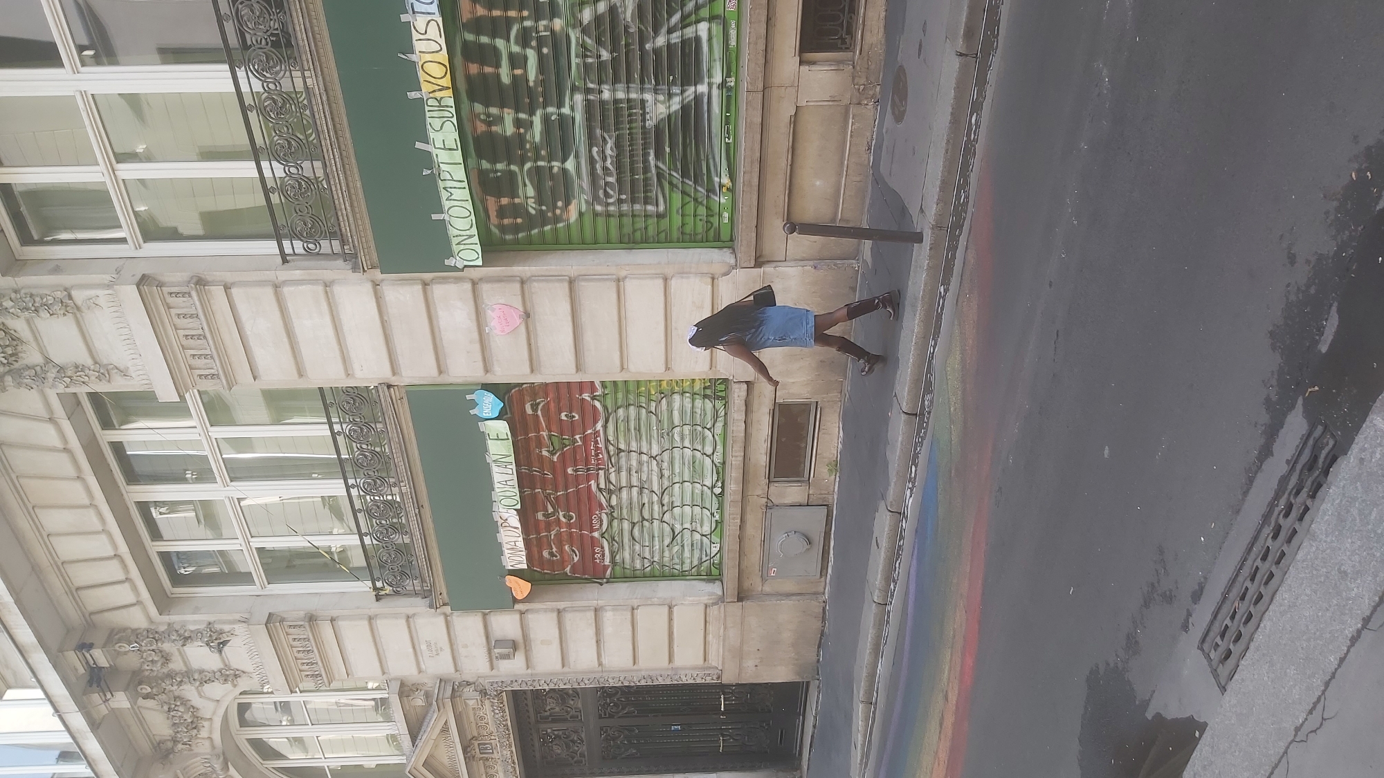 The image depicts a street scene in Paris, France, on a sidewalk in front of a building with ornate architectural details. A Black woman with long, dark hair, wearing a denim dress and brown boots, walks along the sidewalk. She is carrying a black bag over her right shoulder. The building has large windows and storefronts with roll-down shutters. One shutter is covered in red and green graffiti, with text in French such as "Noma dps jouia lan e" and "Ensemble" visible. Another shutter has green graffiti that is less legible. Colorful chalk or paint, resembling a rainbow, is spread across the street and sidewalk in the foreground. A metal drain grate is visible on the street. The day appears to be overcast.