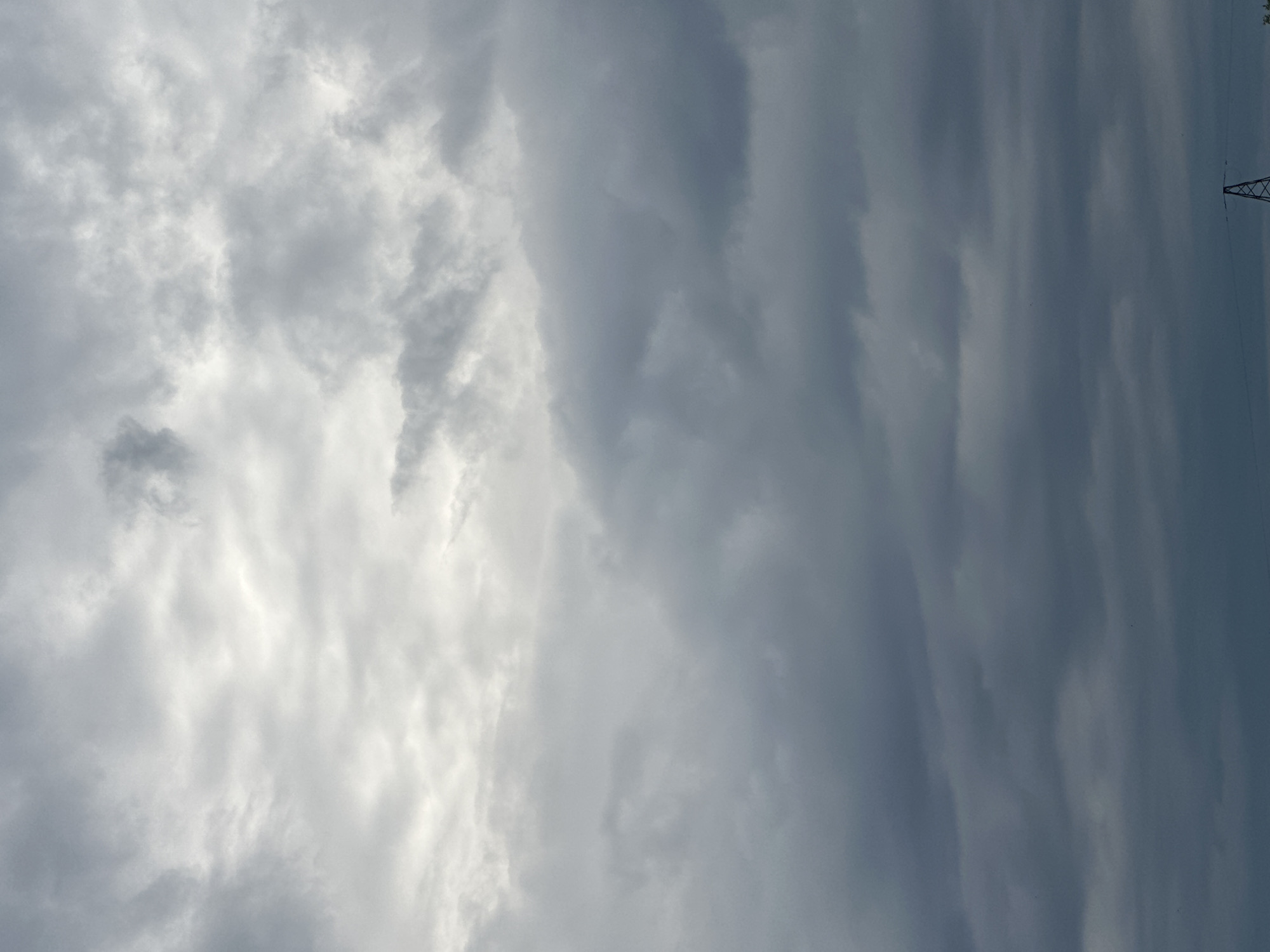 The image depicts a sky filled with various types of clouds. On the left side, there are brighter, more diffused clouds, suggesting sunlight filtering through. As the view moves towards the right, the clouds become darker and more defined, appearing as heavy, layered formations. The overall impression is one of an overcast or changing weather day.
In the far upper right corner, a portion of a tall, metallic pylon or transmission tower is visible against the sky. This suggests an outdoor, possibly rural or semi-rural, setting where such structures are common. There are no people or specific objects apart from the pylon that are clearly discernible. The scene appears to be static, with no discernible activity taking place other than the natural movement of clouds. The lighting and cloud formations do not provide strong cues for a precise time of day, but the ambient light suggests it is daytime. The weather appears to be cloudy, with the potential for rain or unsettled conditions due to the darker, heavier clouds. No text is visible in the image.