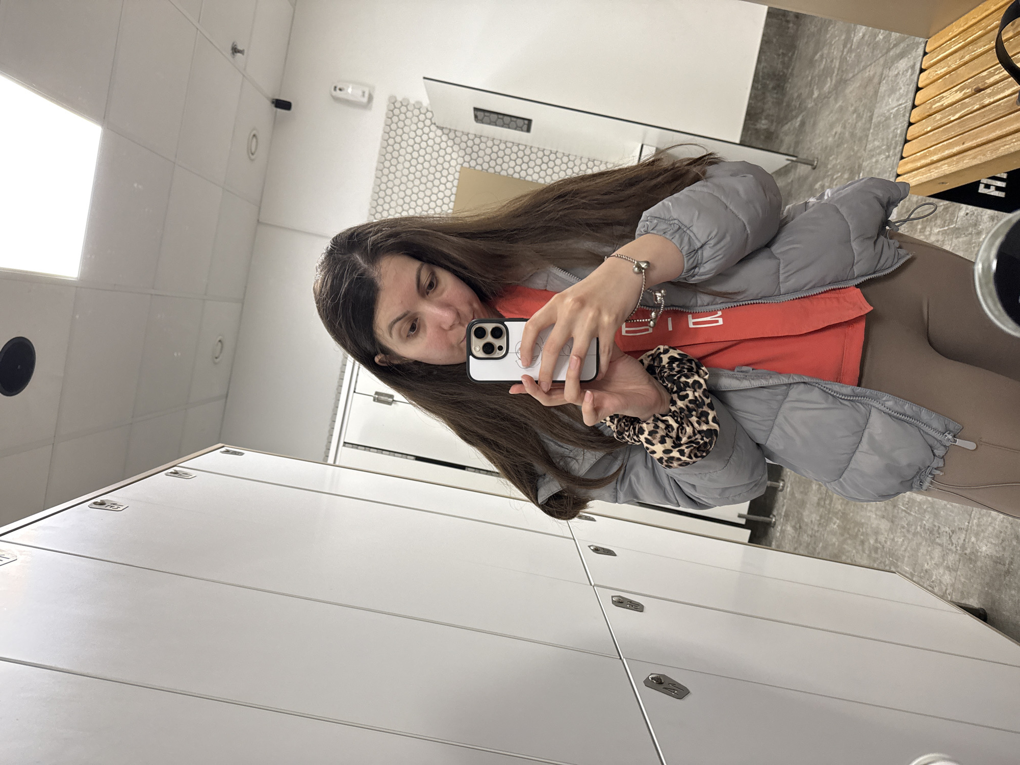 A woman is taking a selfie in what appears to be a locker room. She is holding a white iPhone and looking at the screen. She is wearing a gray puffer jacket over an orange shirt and brown leggings. Her brown hair is long and loose, and she has a leopard print scrunchie on her wrist. The locker room has white lockers and a gray tiled floor. There is a wooden bench in the background, along with a white wall with hexagon tiles.