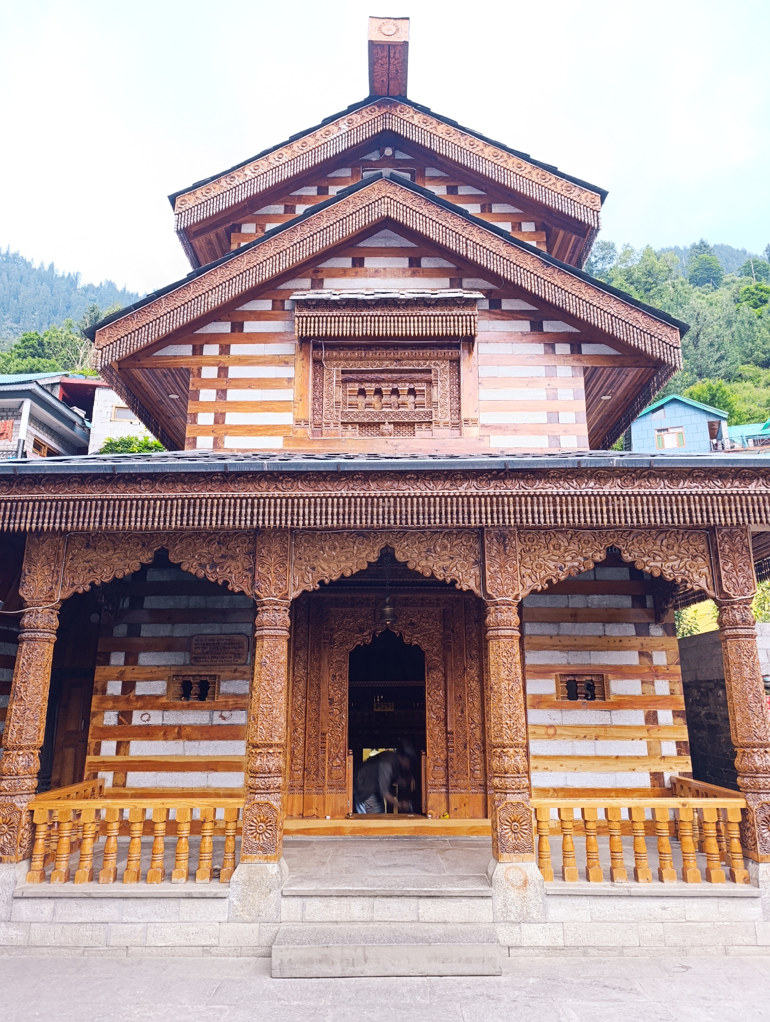 The image shows the exterior of a wooden temple in Vashisht, India. The temple features intricate carvings on its wooden pillars, arches, and rooflines. The structure is made of stone and wood, with the stone walls having horizontal wooden beams. A wooden railing with vertical spindles lines the entrance. The temple is situated in a hilly, green environment, with trees and mountains visible in the background. The sky is overcast. A person, dressed in dark clothing, is partially visible entering the dark doorway of the temple. There is a plaque on the wall to the right of the entrance. Other buildings, including a blue one, are visible behind the temple.