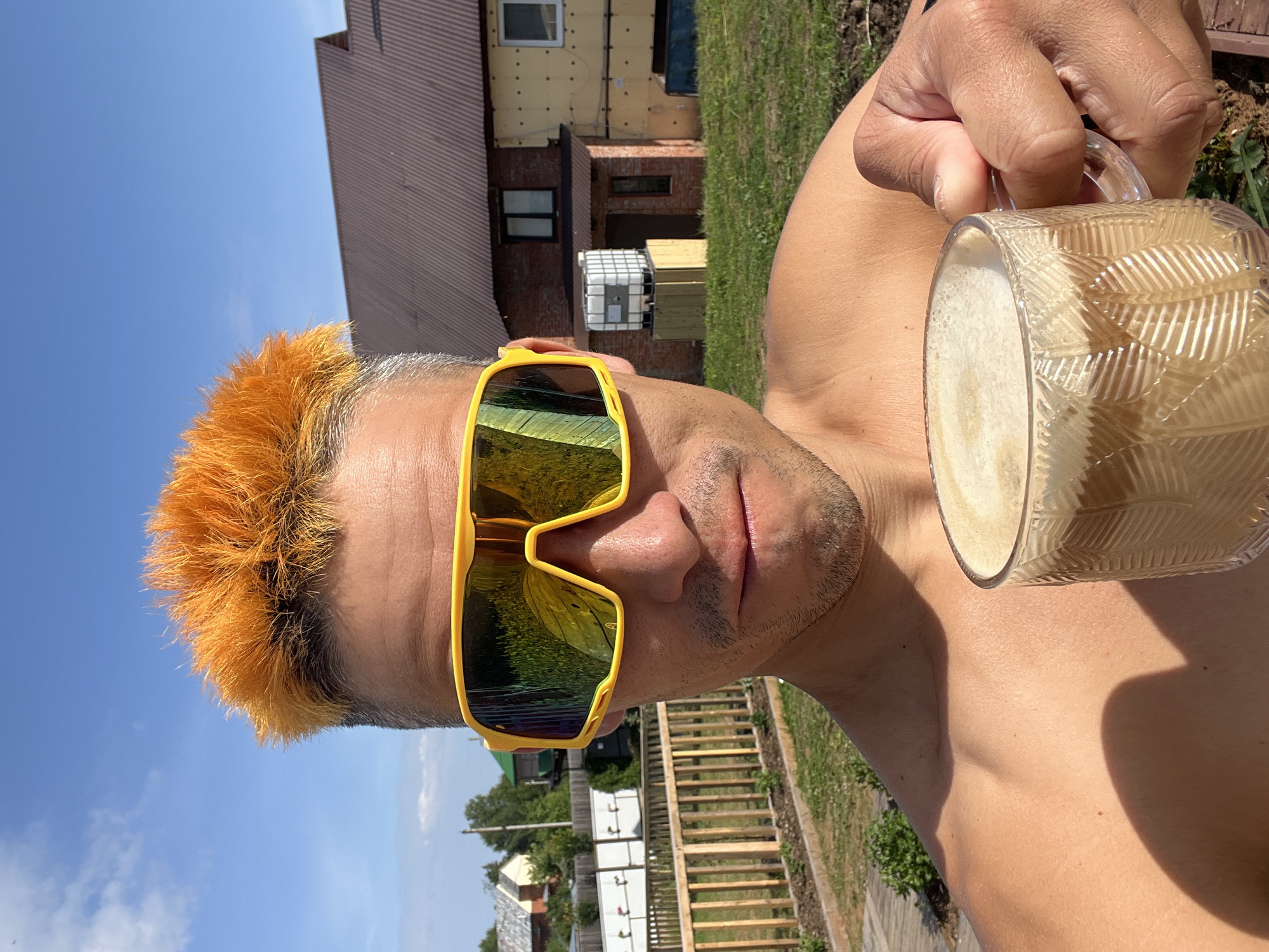 A person with bright orange spiky hair and yellow wraparound sunglasses holds a clear glass mug of coffee with frothed milk. The person is shirtless and appears to be outdoors, with a blurry background of a house, wooden fence, and greenery under a bright blue sky with scattered clouds. The reflection in the sunglasses shows a green, grassy area. The setting suggests a residential area, possibly a dacha or private home in a rural or suburban location, such as Chaikovsky, Russia. The person is looking directly at the camera, holding the mug up in a selfie-style pose. The sunlight is strong, casting shadows on the person's chest.