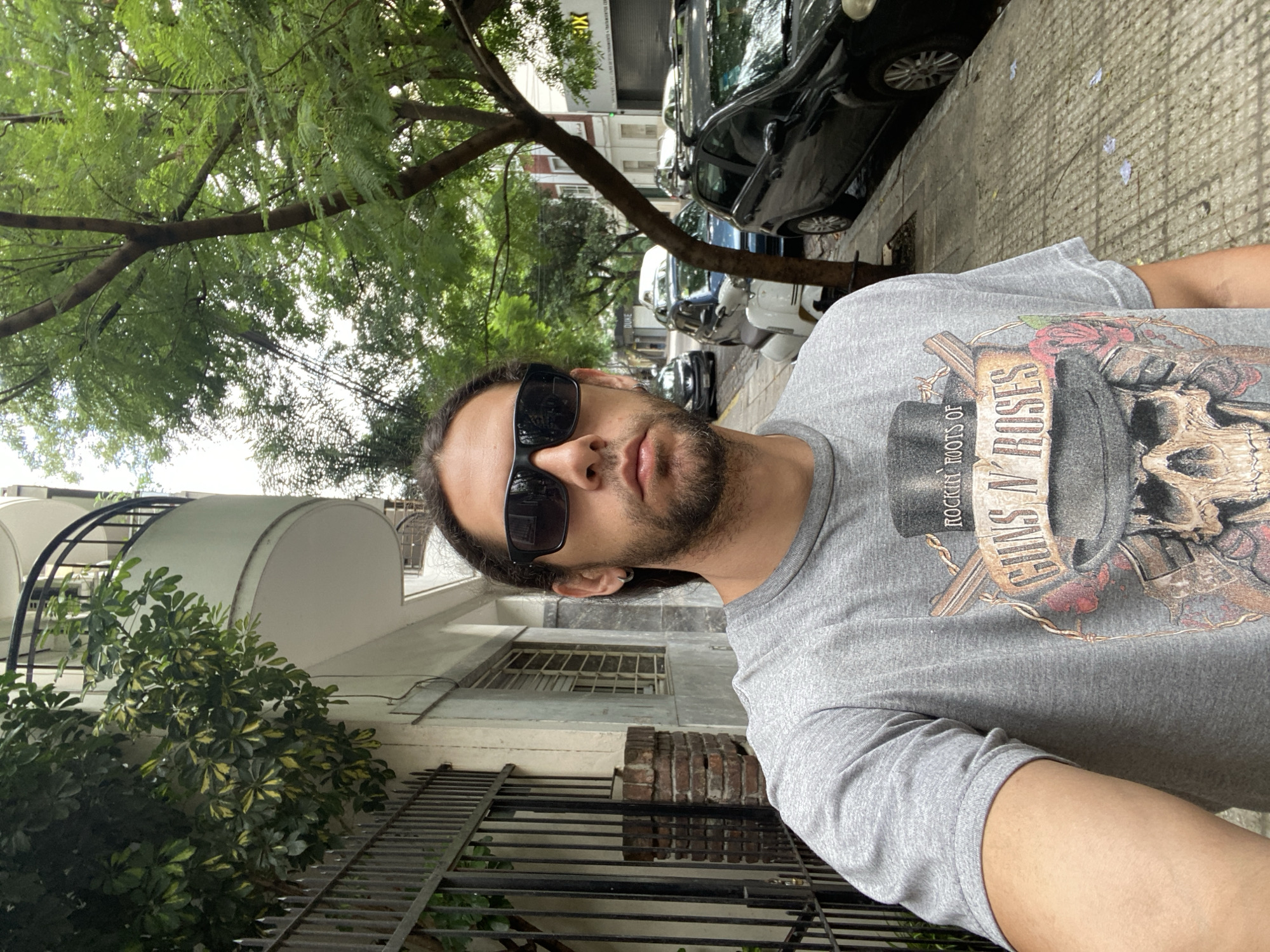 A man with sunglasses and a Guns N' Roses t-shirt is taking a selfie on a sidewalk. He has dark hair pulled back and a beard. Behind him is a building with a rounded balcony and black iron railing. A tree partially obscures the building. Further down the street are several parked cars. The street appears to be located in a residential area. Based on the architecture and the presence of certain types of trees, the location is likely in Buenos Aires, Argentina.