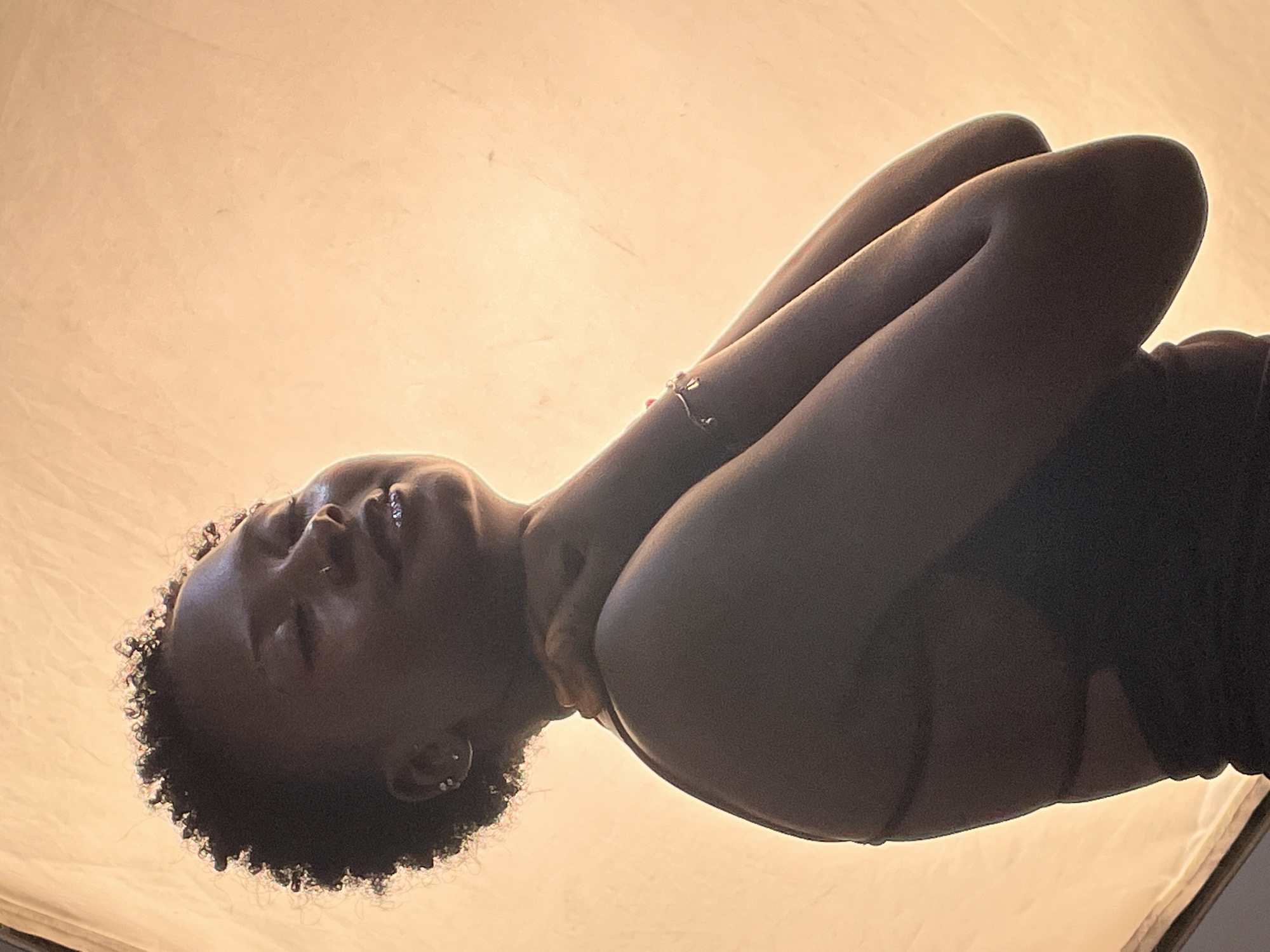 A medium shot captures a Black woman with short curly hair, positioned against a backdrop of light yellow. Her arms are crossed above her head. She appears to be wearing a dark sleeveless shirt. There is no indication of the city of Jos, Nigeria.