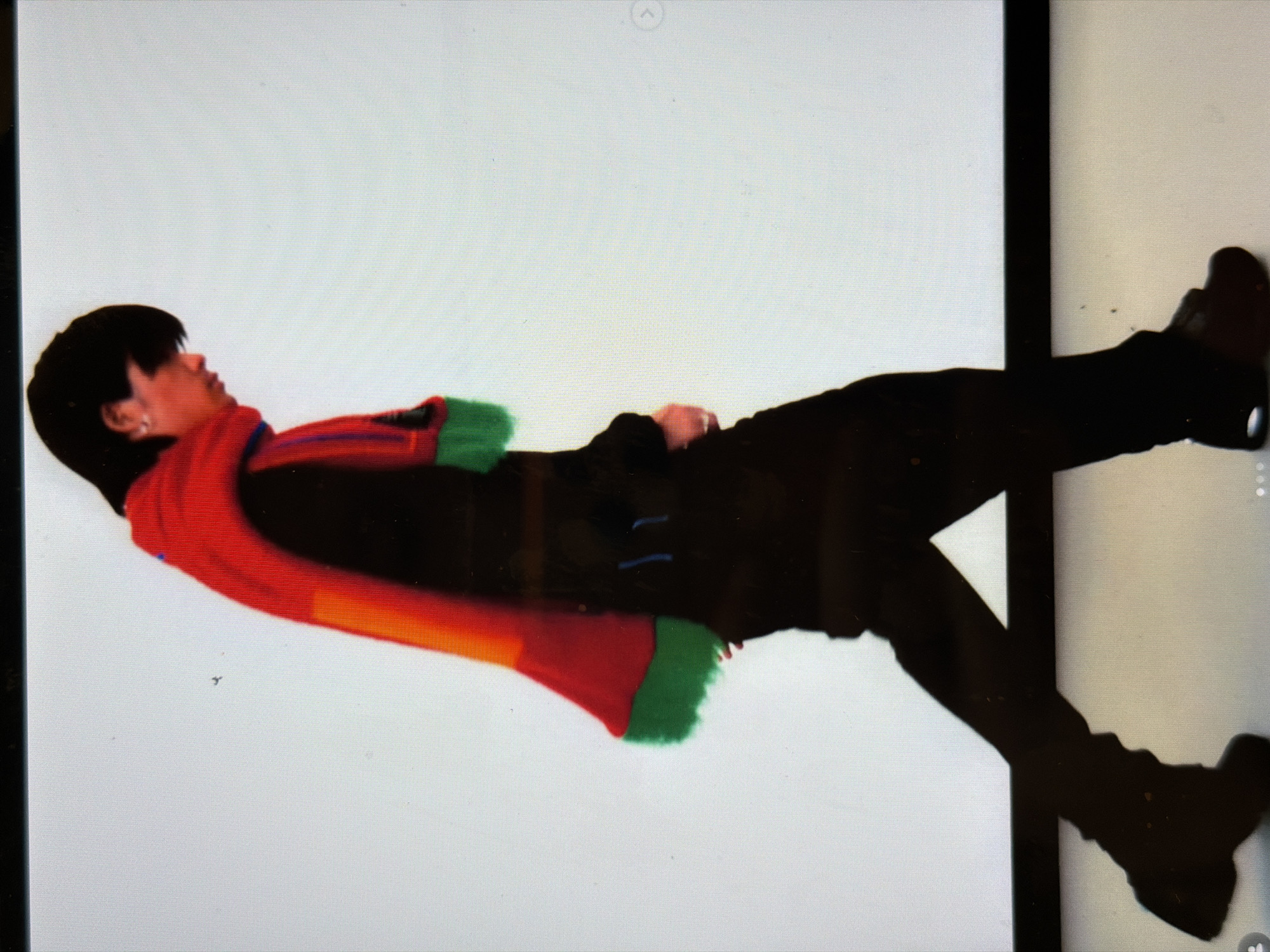 The image shows a person lying down against a white wall or backdrop. The person has dark hair and is wearing a dark-colored jacket. A red, orange, and green scarf is draped around their neck. The person's legs are extended and slightly crossed, and they are wearing dark-colored shoes. A black vertical line is visible on the right, possibly indicating a door frame or the edge of a wall. The location is not discernible from the visual information provided.