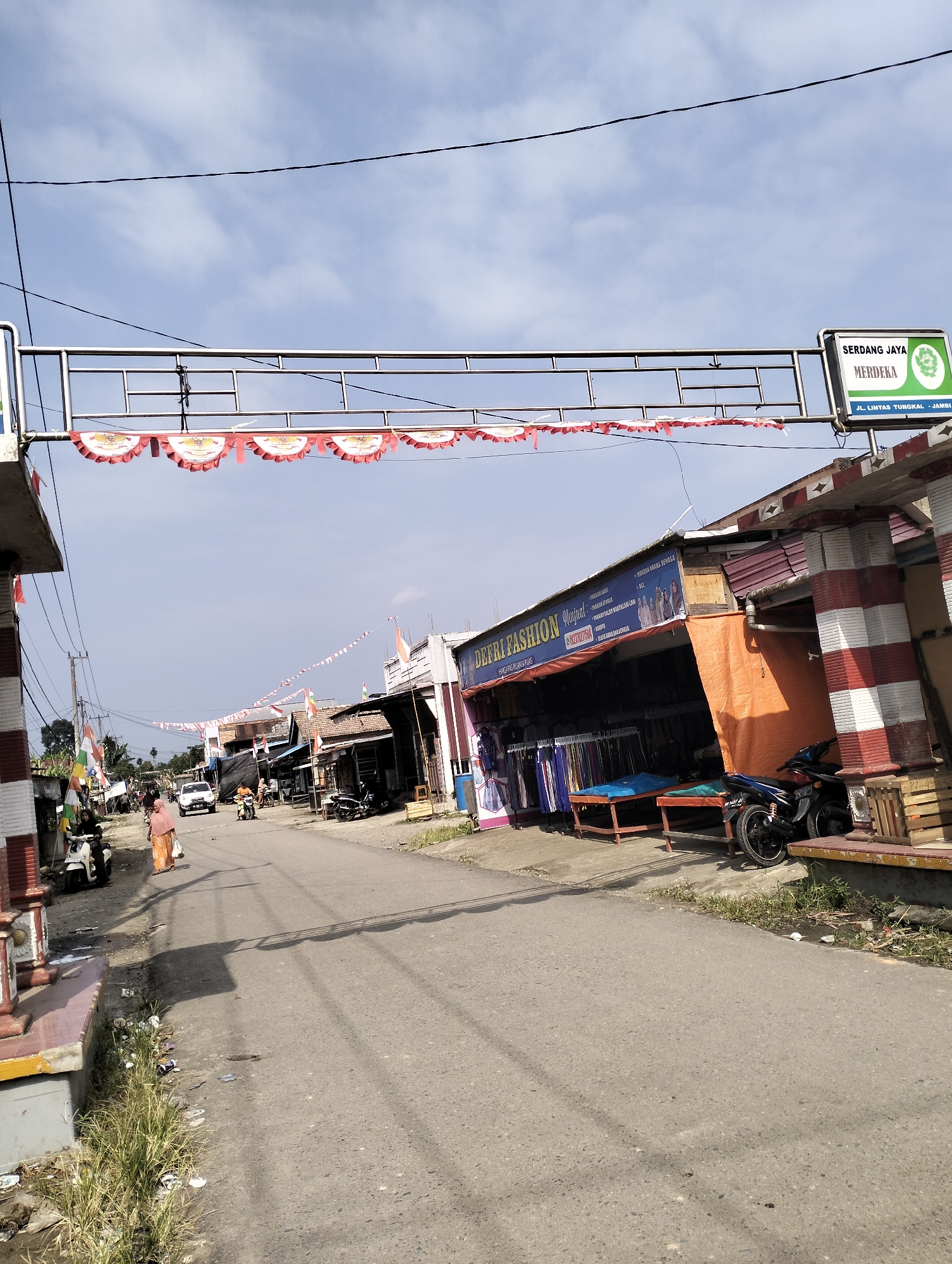 The image depicts a street scene in Pematang Lumut, Indonesia, likely a commercial area with several small shops lining the road. An elevated metal archway adorned with red and white festive banners, possibly for a national holiday, spans across the street. A sign on the right side of the archway reads "SERDANG JAYA MERDEKA" and below it, "JL. LINTAS TUNGKAL - JAMBI".
Several people are visible on the street. A woman wearing a light orange headscarf and an orange patterned skirt walks on the left side of the road. Two individuals are near a white scooter on the far left. Further down the street, other pedestrians and several motorcycles are present, some parked and some in motion. Vehicles, including a white car, are also visible on the road.
The shops on the right have open fronts displaying merchandise, primarily clothing. One shop sign reads "DEFRI FASHION". Orange tarps are used as awnings for some of the stalls. The street itself is paved with asphalt and shows some debris and vegetation growing along its edges. Power lines are strung across the sky, typical of urban or semi-urban environments. The sky is partly cloudy. The overall impression is that of a bustling local market or commercial strip.