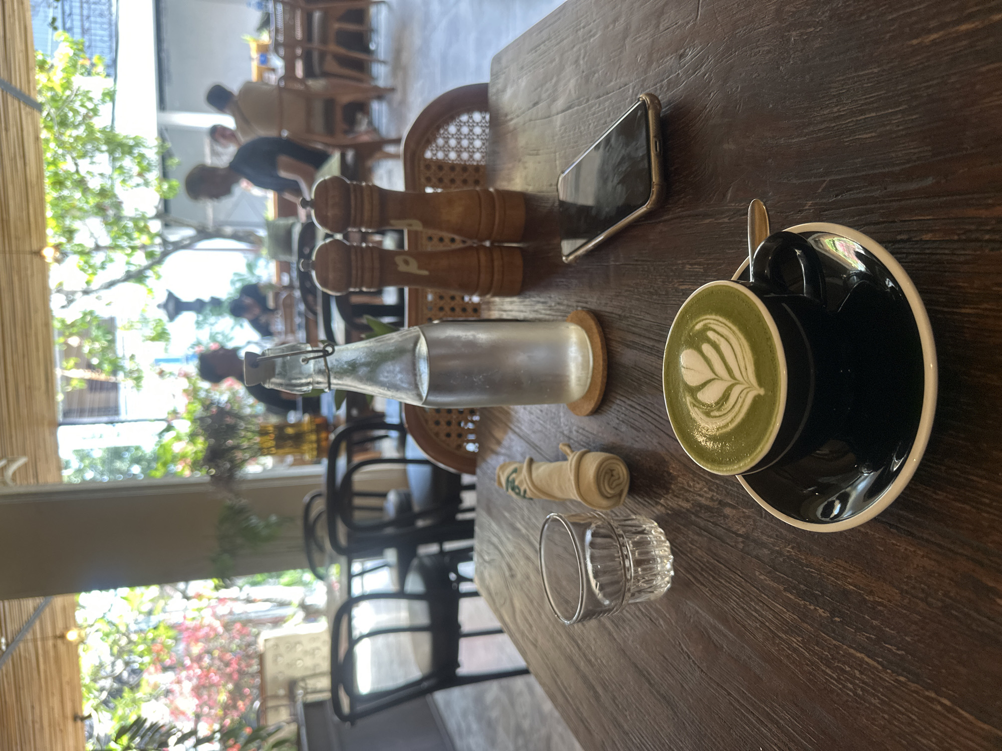 The image shows a close-up view of a dark wooden table in a cafe. In the foreground, there is a black cup and saucer containing a matcha latte with latte art. To the left of the latte is a small glass of water, a rolled-up napkin, and a bottle of water with a stopper. Further back on the table are two wooden salt and pepper grinders. A smartphone is placed face down to the right of the grinders. In the background, the cafe is visible with several tables and chairs. People are seated at some of the tables, and lush green foliage can be seen through large windows. The overall ambiance suggests a relaxed and modern cafe setting.