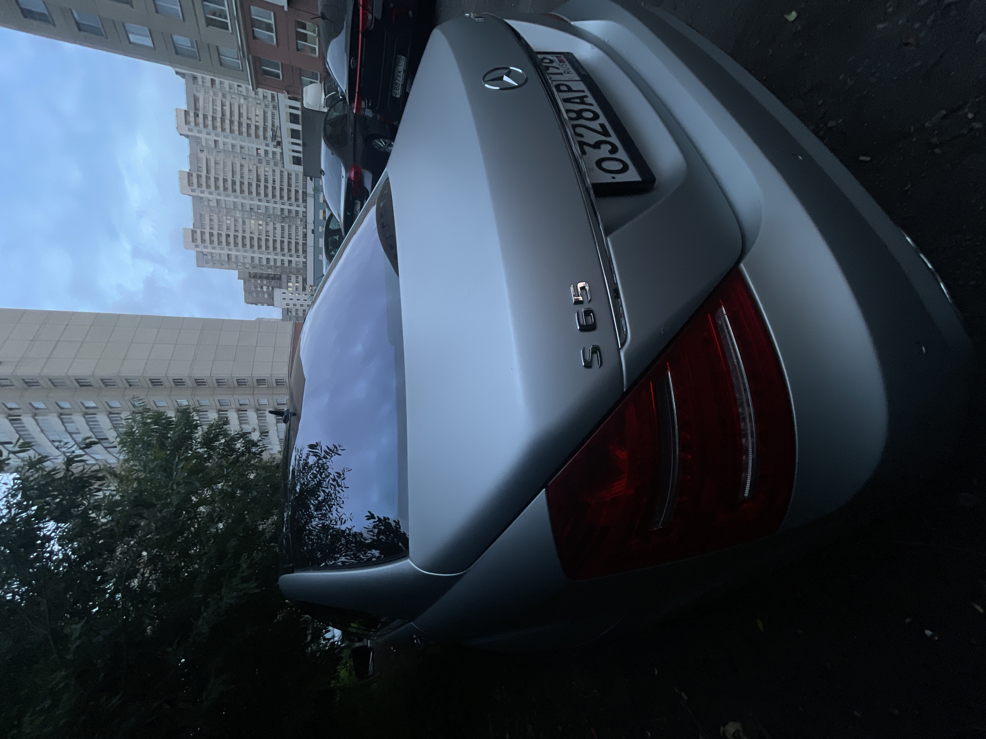A silver Mercedes-Benz S65 is parked outdoors in what appears to be an urban setting. The car is the primary subject, seen from a rear-quarter angle, with its taillight and rear bumper visible. The license plate is partially obscured but reads "0328AP 178 RUS". In the background, tall apartment buildings and trees are present, under a cloudy sky. Other cars are parked in the vicinity, including a dark sedan and a Kia. The lighting suggests it is either early morning or late evening.