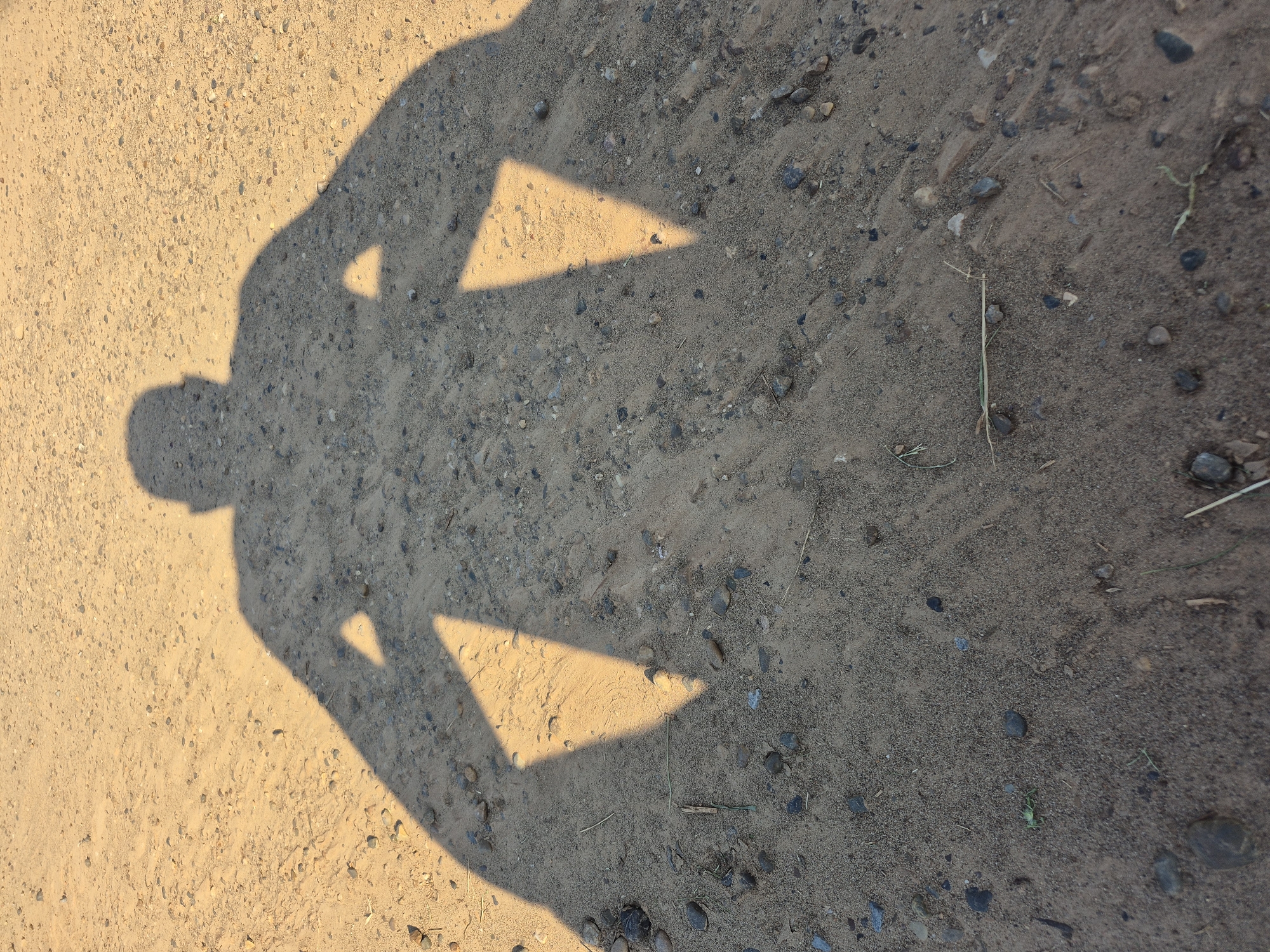A long, dark shadow of a person stands on a sandy path. The shadow is cast by the sun, which appears to be low in the sky, judging by the length of the shadow. The path is made up of sand and small pebbles, and there are a few dry blades of grass scattered across it. The shadow itself is that of a person with their arms outstretched to the sides. The head of the shadow is rounded, and the body appears to be broad-shouldered. The arms are extended outwards, and there are two triangular shapes of light within the shadow of each arm, suggesting the sun is shining through gaps or openings in the person's clothing or a held object. The scene is illuminated by warm, golden sunlight.