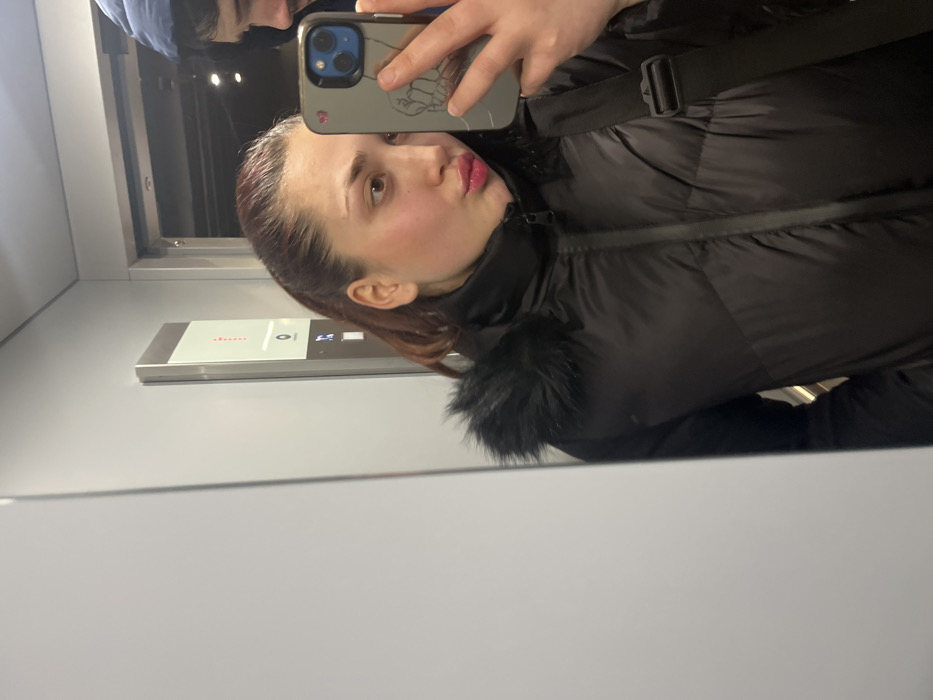 A young woman is captured in a mirror selfie taken inside an elevator. She is the central subject, positioned sideways in the frame, wearing a black puffer jacket with a faux fur collar and a black shoulder bag. Her auburn hair is pulled back, and she is looking towards the camera (reflected in the phone), with her lips slightly puckered in a playful 'duck face' expression. She holds a smartphone with a gray case, which has a simple line drawing on its back.
Partially reflected behind her, another individual's head and shoulder are visible, wearing a blue hooded garment. The setting is an elevator cabin, characterized by light gray walls and the reflective surface where the photograph is taken. To the woman's right, an elevator control panel is visible, featuring a display showing a red dotted pattern, a button labeled "0", and illuminated icons for an up arrow, down arrow, a bell, and door open/close functions. The overall scene suggests a casual moment, possibly while waiting or traveling in an elevator in Schiltigheim, France. The lighting is artificial and internal to the elevator.