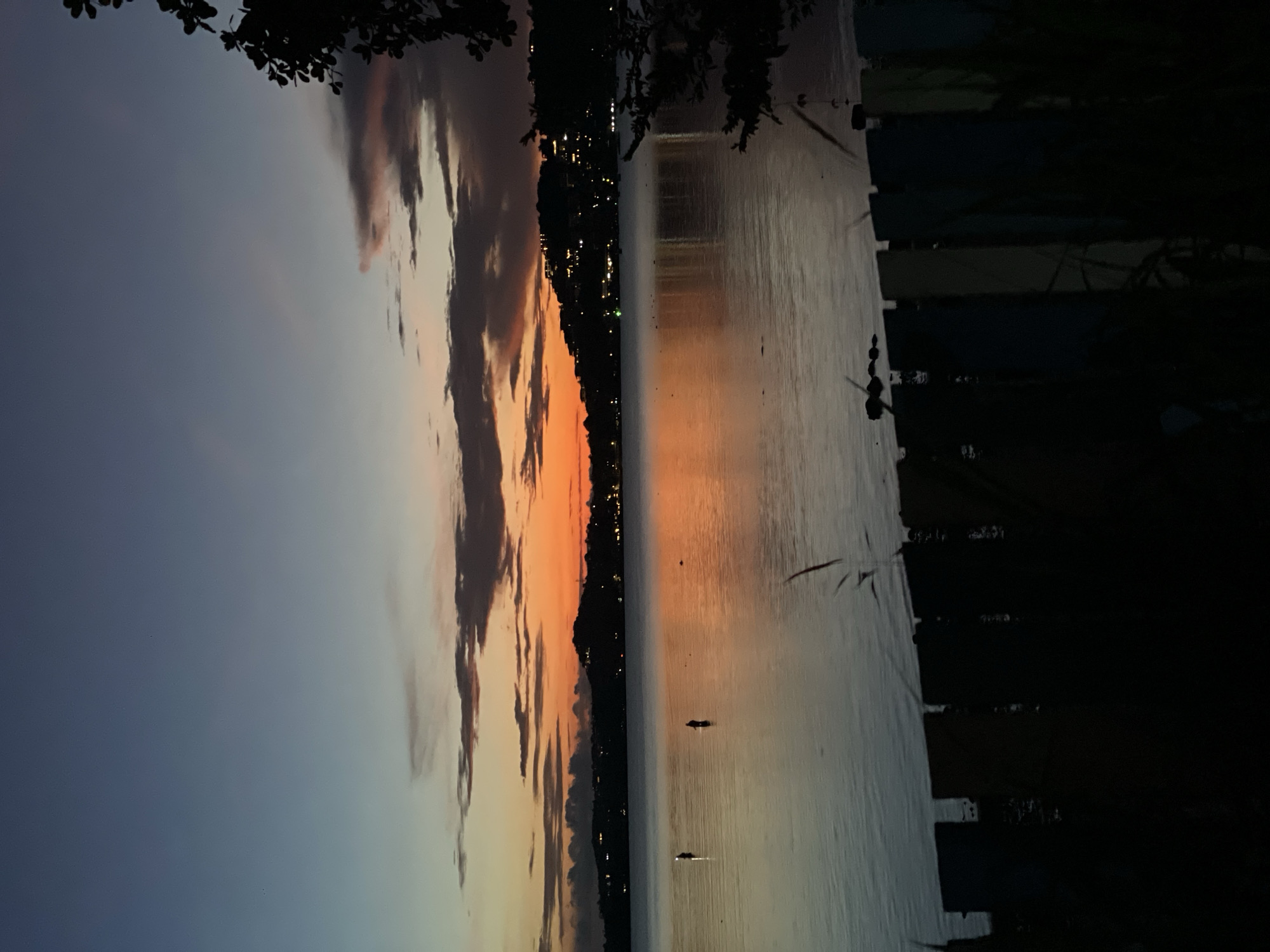 This image depicts a sunset over the sea in Baan Bang Rak, Thailand. The sky transitions from a deep blue at the top to vibrant orange and yellow hues near the horizon, with scattered clouds adding texture. In the water, two figures, likely people, are visible standing in the shallows, possibly fishing. The distant shoreline is lined with trees and dotted with lights from buildings, suggesting a populated area. In the foreground, a dark silhouette of a fence and some vegetation obscures the lower part of the scene. The water reflects the colors of the sky, creating a serene and tranquil atmosphere.