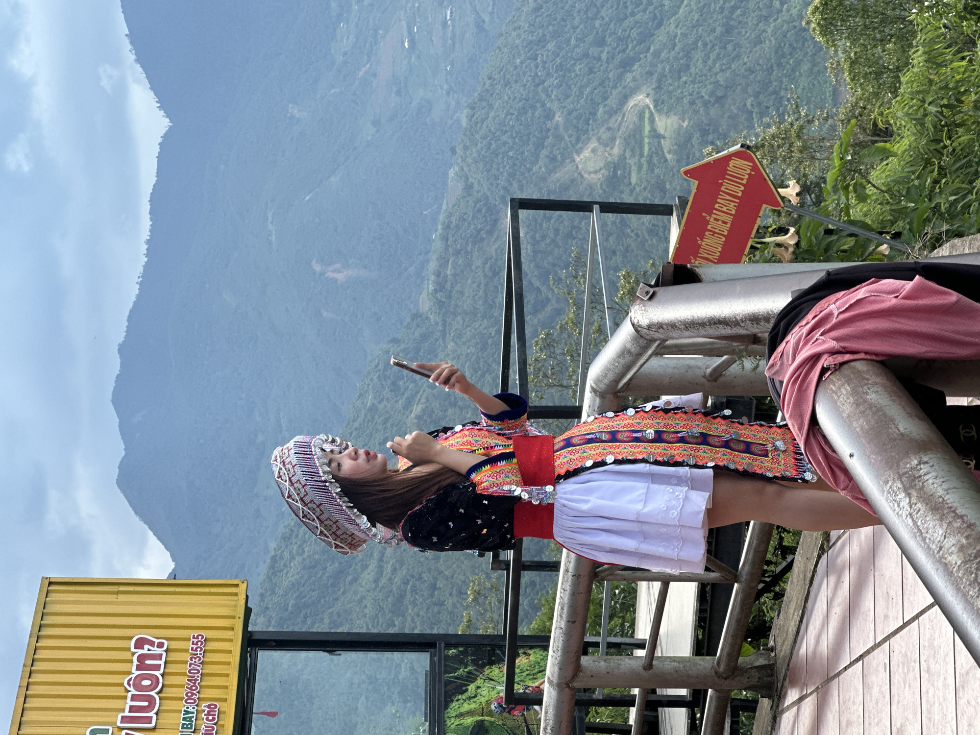 A lone female, dressed in traditional attire, stands on a tiled viewing platform overlooking a vast mountainous landscape in Tu Le, Vietnam. She has long dark hair and wears an elaborate headpiece adorned with silver-colored beads and red/white patterns. Her ensemble includes a black long-sleeved top with embroidered elements, a colorful tunic featuring intricate patterns in orange, red, and blue, embellished with coin-like silver discs, a wide red sash around her waist, and a white pleated skirt. In her left hand, she holds a silver-colored smartphone, looking at its screen. Her right hand is raised near her mouth, possibly applying a cosmetic product.
The background consists of multiple layers of lush green, cloud-covered mountains under an overcast sky. A winding dirt path is visible on one of the distant slopes. To the left, a yellow sign, partially visible, displays Vietnamese text "luôn?", "BAY: 0964.073.555", and "CHỖ", suggesting a contact number for a flying activity. In the lower right foreground, a red arrow-shaped sign with white text points right, reading "LỐI XUỐNG ĐIỂM BAY DÙ LƯỢN", which translates to "PATH DOWN TO PARAGLIDING POINT", confirming the location as a paragliding site. Metal railings, showing signs of wear, border the platform. A pink garment and a black garment are draped over a railing section in the immediate foreground. Several light-colored bell-shaped flowers are visible near the red sign.