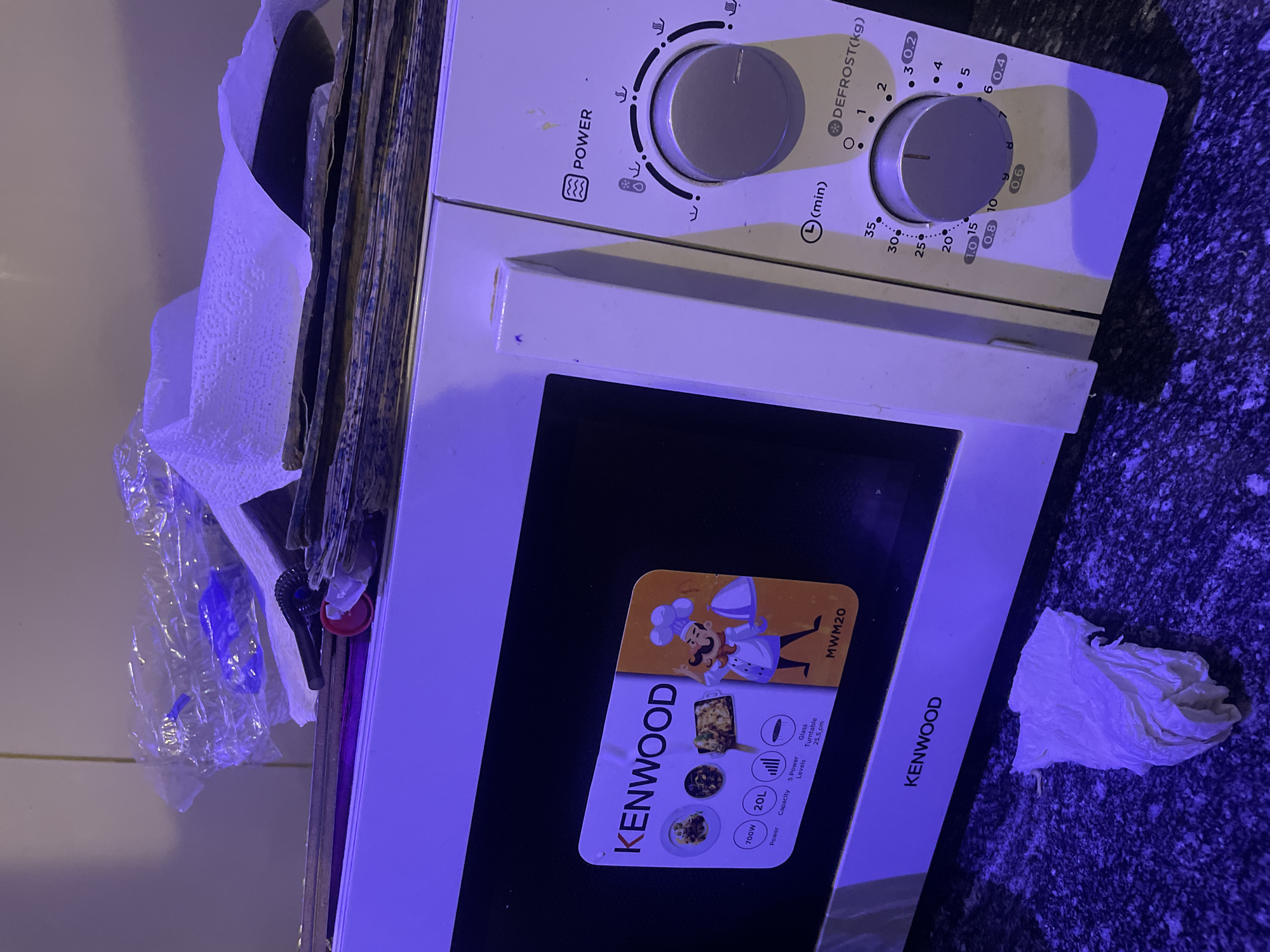 The image shows a white Kenwood microwave oven on a dark speckled countertop. On top of the microwave, there is a stack of papers, some plastic wrap, and crumpled paper towels. To the left of the microwave, on the countertop, is a crumpled paper towel. The microwave has a black door with a Kenwood logo and a sticker detailing its specifications: 700W power, 20L capacity, 5 power levels, and a 25.5 cm glass turntable. The control panel on the right side of the microwave features a power setting, a timer dial marked in minutes and with settings for defrosting in kilograms, and various icons indicating cooking functions. The scene is illuminated by purple lighting, casting shadows on the countertop.