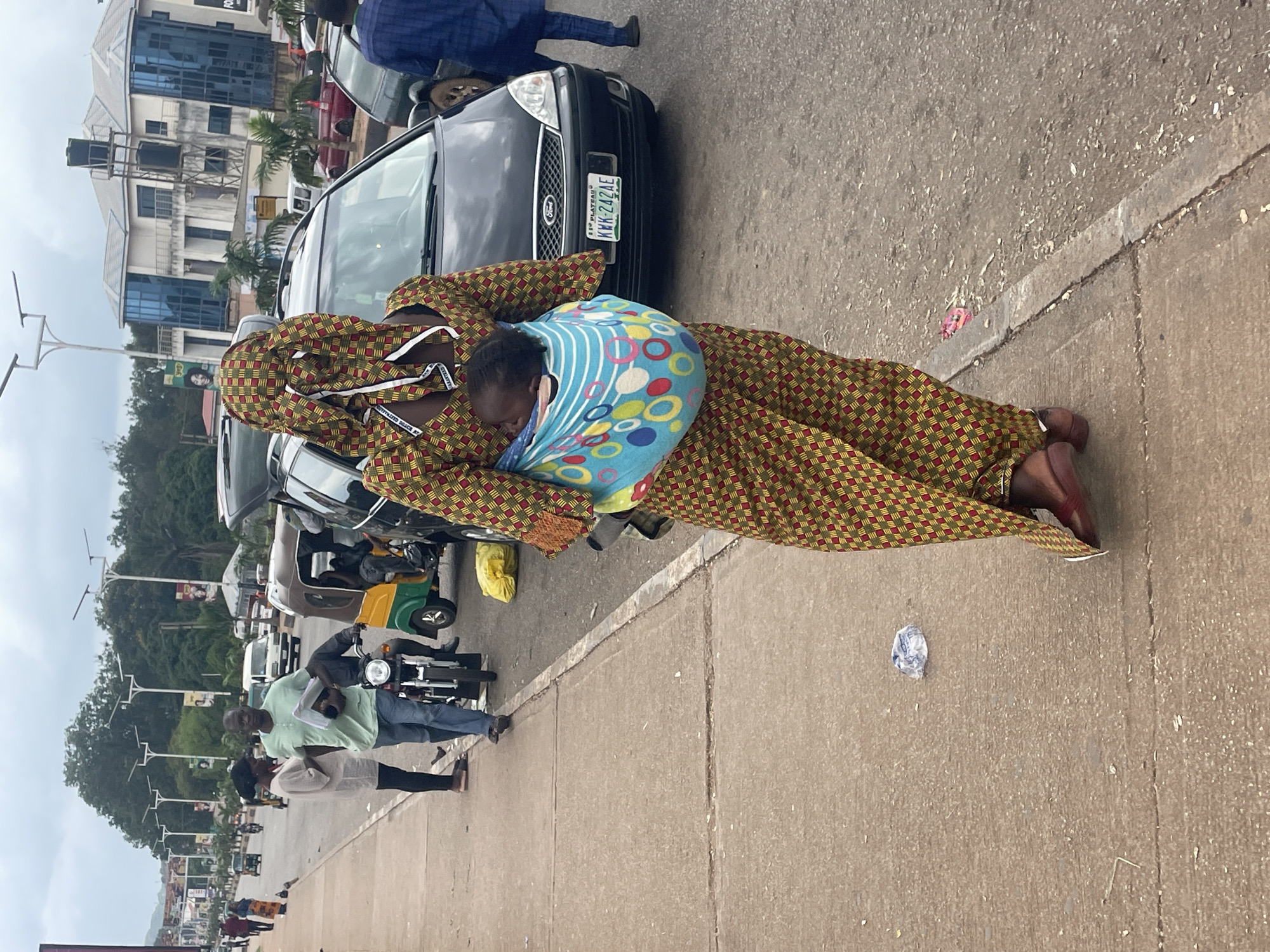 A rear view of a woman wearing a patterned dress and headwrap, carrying a baby in a sling, walks on a concrete sidewalk in Barakin Lerawi, Nigeria. The woman's dress features a geometric pattern of red, yellow, and green squares with thin black lines. The baby, wrapped in a blue and colorful polka-dotted sling, is visible on the woman's back. Behind the woman, a black Ford Focus with license plate KWK-242AE is parked on the street, along with other vehicles including a three-wheeled auto-rickshaw. The street is lined with buildings and trees, and a cloudy sky is visible above. The sidewalk is worn and shows some debris, including a crumpled plastic wrapper.