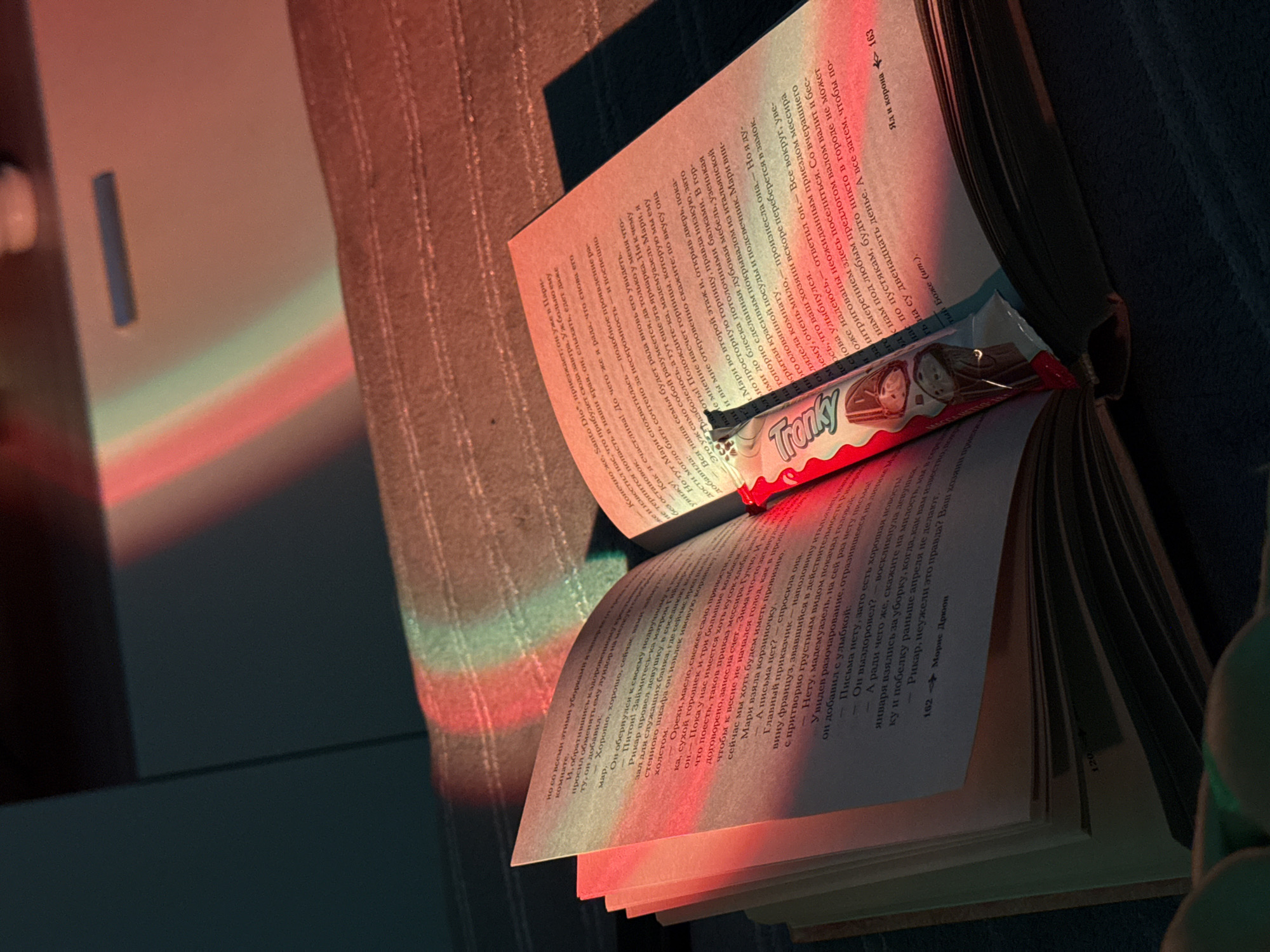 The image captures an open book lying on a soft surface, with a candy bar placed between its pages. The book is in Russian, judging from the script on the pages. The candy bar is a "Tronky" wafer snack. The scene is bathed in a spectrum of colorful light, casting hues of red, green, and blue across the pages and surrounding surface. The setting appears to be indoors, possibly in a home environment. There are no people visible in the image. The focus is on the juxtaposition of the book, the candy, and the colored light, creating a sense of quiet leisure.