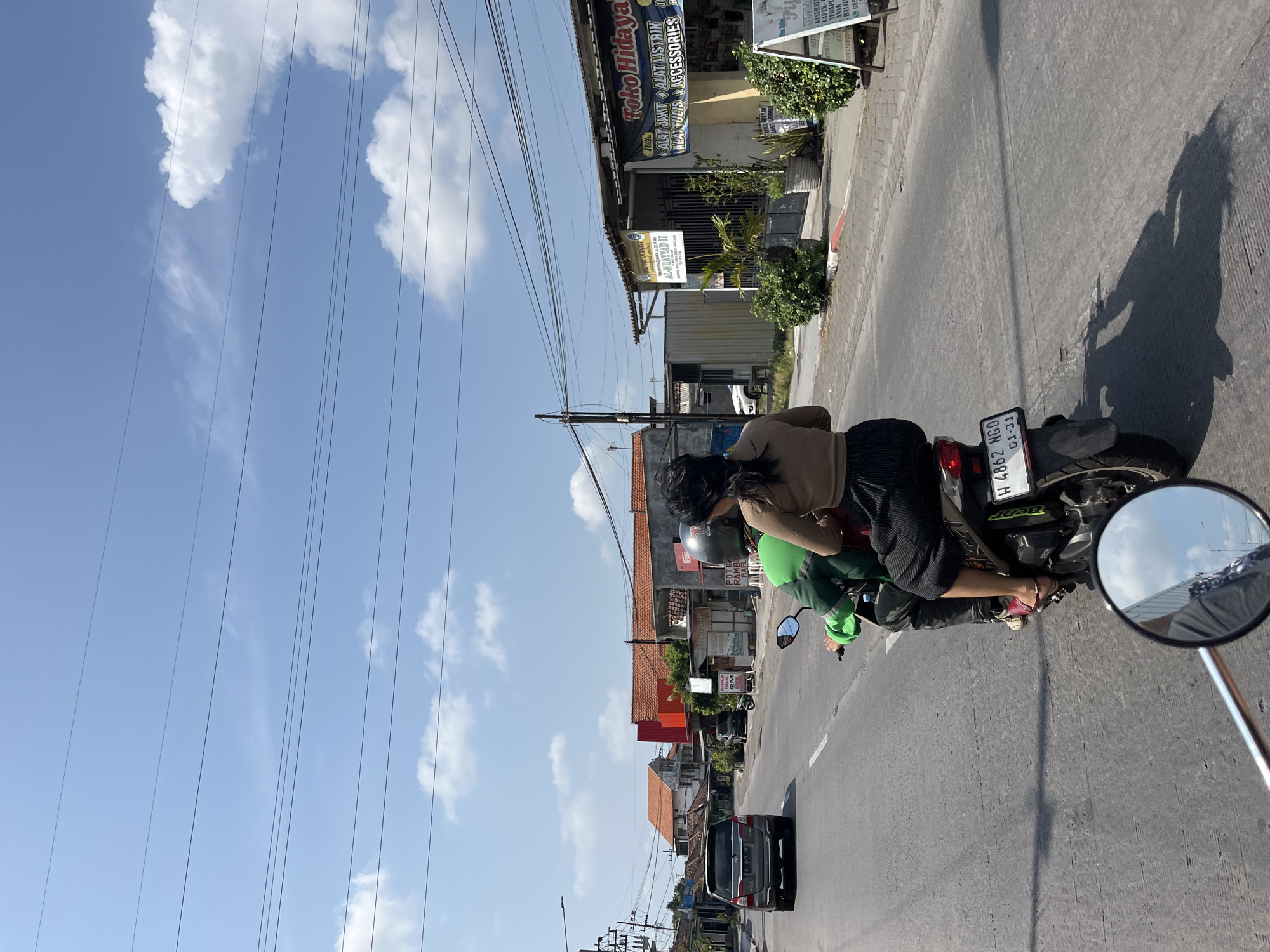 The image depicts a street scene in Boro, Indonesia, with a clear blue sky and scattered white clouds overhead. A paved road runs through the center of the frame, with buildings lining the right side. In the foreground, two people are riding a scooter from the perspective of someone on the same scooter, indicated by the partial reflection in the rearview mirror. The driver wears a green jacket and a helmet, while the passenger, a woman, has dark hair and is wearing a beige top and a black skirt. The scooter has a white license plate with the registration "W 4862 NGO" and "01-21". On the right side of the road, shops are visible, including one with a sign that reads "Toko Hidaya" and another listing "AL-MUAYYAD II". A grey SUV is also visible on the road in the left lane, moving away from the viewer. The environment appears to be a typical urban street with utility poles and overhead wires.