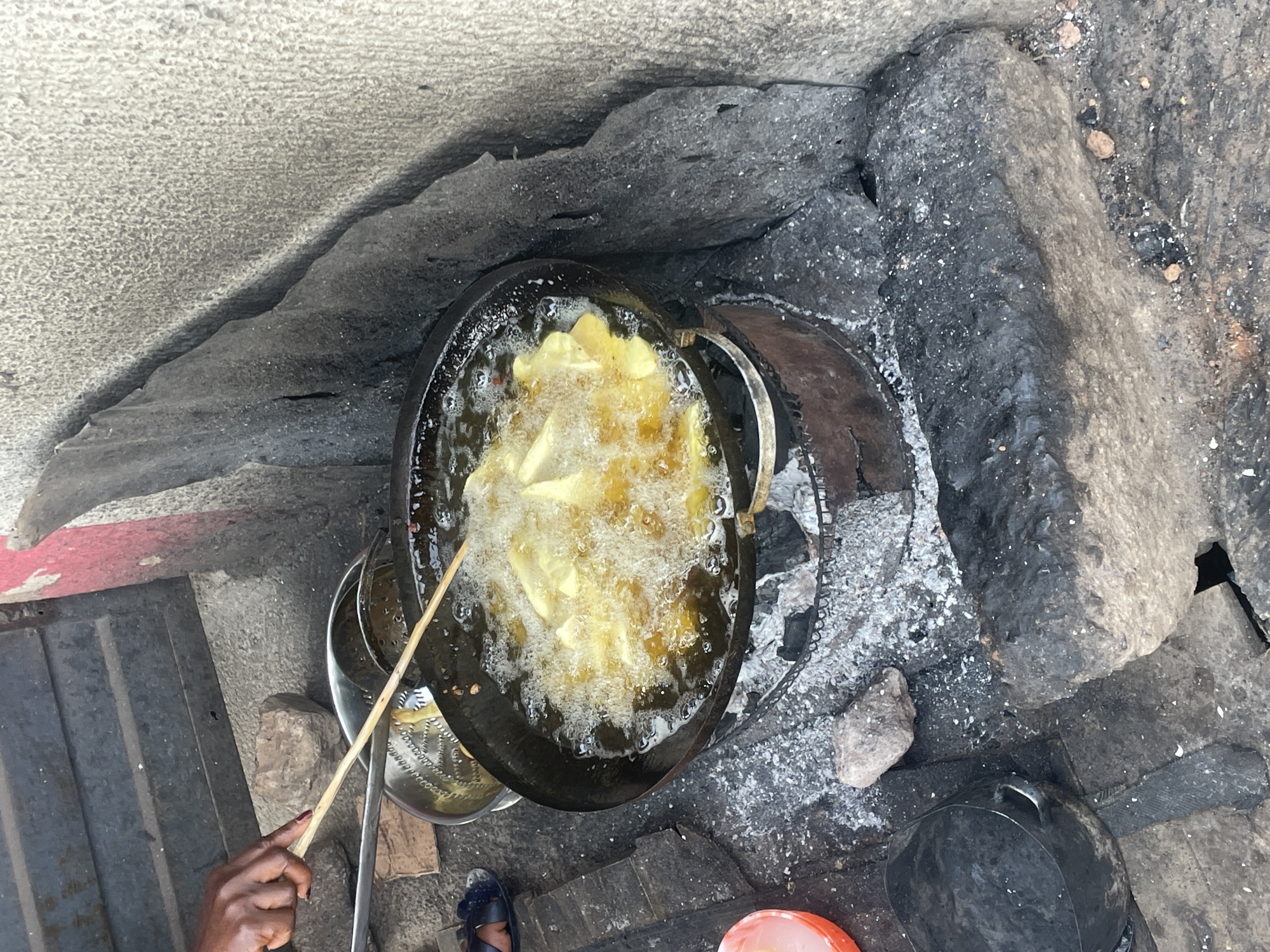 An overhead shot captures an outdoor cooking scene in Jos, Nigeria, centered on a large black pot vigorously frying food, likely chips or yam, in bubbling oil. A dark-skinned hand, with painted fingernails, is visible on the left, holding a long wooden stick used to stir or manage the food in the hot oil.
The cooking pot rests on a makeshift stove composed of large, dark grey rocks, which appear to have been used for an open fire, evidenced by the scattered white ash and black charcoal residue surrounding the pot and on the ground. To the left of the main pot, a metal sieve or strainer sits in a metallic bowl, ready to drain the fried items.
The ground around the cooking area is a mix of concrete or rough stone, with some smaller rocks and debris scattered about. A black cooking pot with a handle and an orange plastic bucket are visible towards the bottom right of the frame. A foot clad in a dark sandal is also visible on the lower left. A red line runs along the edge of the ground or a nearby structure on the left side of the image. The overall setting suggests a bustling, functional outdoor food preparation area, common for street food or communal cooking. No specific time of day or weather conditions can be determined, and no text is visible.