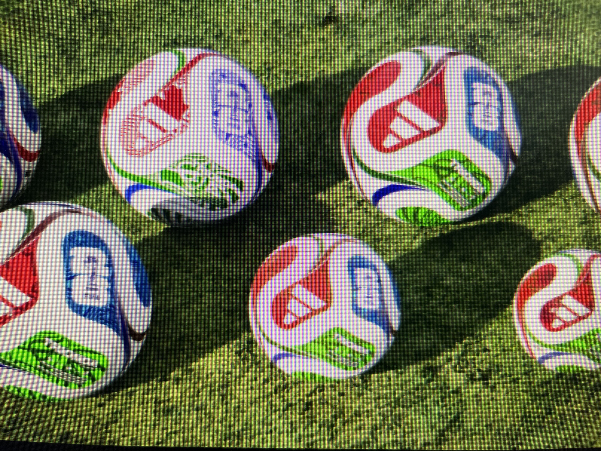 The image shows six Adidas soccer balls on a green, textured surface resembling artificial turf. The balls have a white base with red, blue, and green abstract patterns and logos. Some of the balls feature the Adidas logo and what appears to be the FIFA World Cup trophy logo. The lighting suggests an outdoor setting, possibly a sports field. There are no people present in the image. The image does not contain any specific landmarks or elements that would allow for the identification of the city of Varna, Bulgaria.