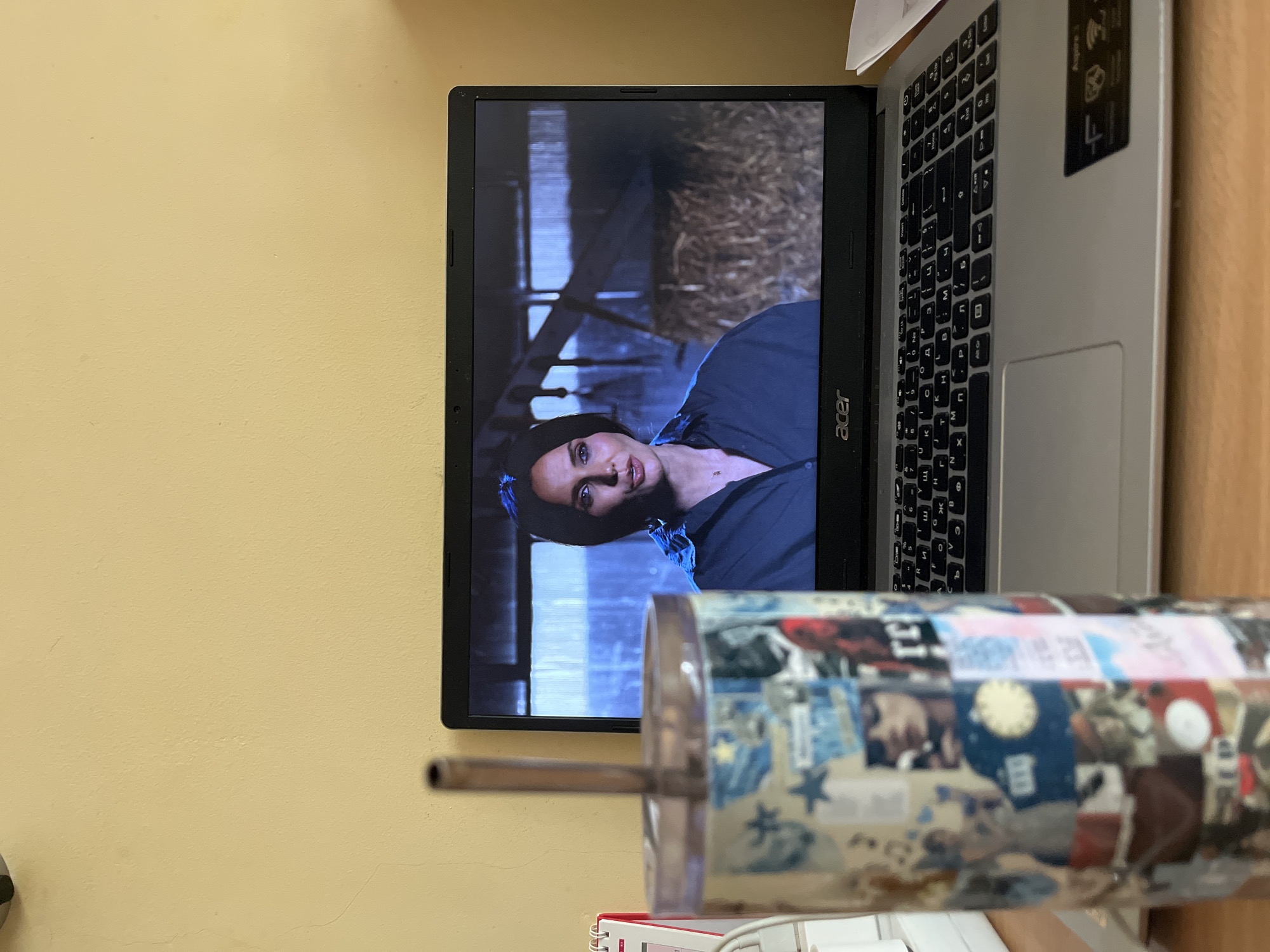 A laptop displaying a scene with Angelina Jolie in the movie "Those Who Wish Me Dead" is on a wooden desk next to a decorated drinking cup with a straw. The background is a pale yellow wall. The image was likely taken indoors. No location information related to Sofia, Bulgaria is discernible in the image.