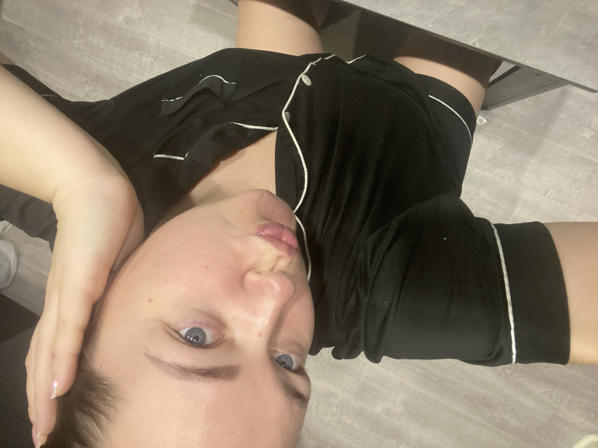 An adult female is depicted in a high-angle, overhead selfie, looking up towards the camera with her head tilted to the left. She has fair skin, blue eyes, and light brown hair, which appears pulled back. Her lips are pursed, and a slight flush is visible on her cheeks. Her right hand rests on the top of her head, with manicured fingernails visible. She is wearing a black short-sleeved top featuring white piping along the collar and placket, secured by white buttons. Matching black shorts with white piping are also visible on her lower body. The immediate background consists of light grey or light wood-look laminate flooring, and a dark, reflective surface, possibly a piece of furniture, is visible on the right side of the frame. The specific location in the city of Sochi, Russia, or any other precise geographical location, cannot be determined from the visual information provided in the image.