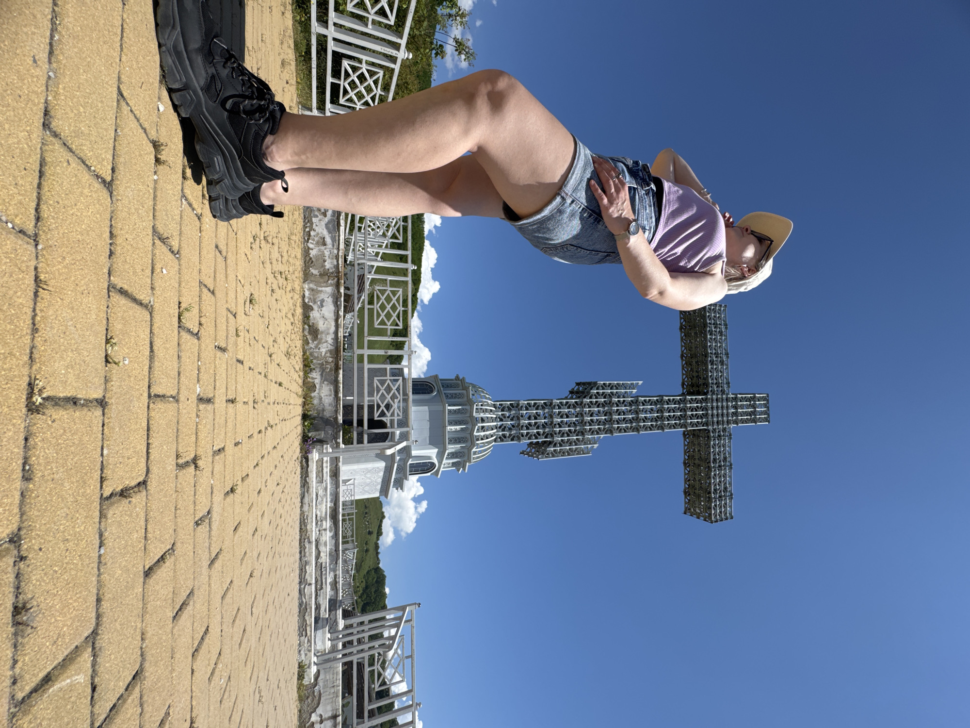 A bright, sunlit outdoor scene in Gelendzhik, Russia, features a person standing on a paved walkway overlooking a vast blue sky with a significant monumental structure. The image, originally rotated, shows a person, likely female, positioned in the foreground on yellowish-brown interlocking paver stones. She is wearing chunky black sneakers, light denim shorts, and a light purple ribbed tank top. A light-colored straw hat, dark sunglasses, a watch on her left wrist, and rings on her fingers complete her attire. Her posture is relaxed, with one hand casually placed on her hip, suggesting she is observing the view or posing for a picture.
In the mid-ground and background, against a clear, vibrant blue sky with a few scattered white clouds, stands a massive, metallic lattice-work cross monument. To the left of the cross, an ornate, light-colored building with a domed roof and decorative arches is visible, possibly a chapel or part of the monument complex. White metal railings with intricate geometric patterns line the paved area, separating it from the scenic background. Beyond these railings, distant green hills and trees are visible, indicating an elevated viewpoint. The weather is clear and sunny, indicative of daytime. There is no visible text in the image.