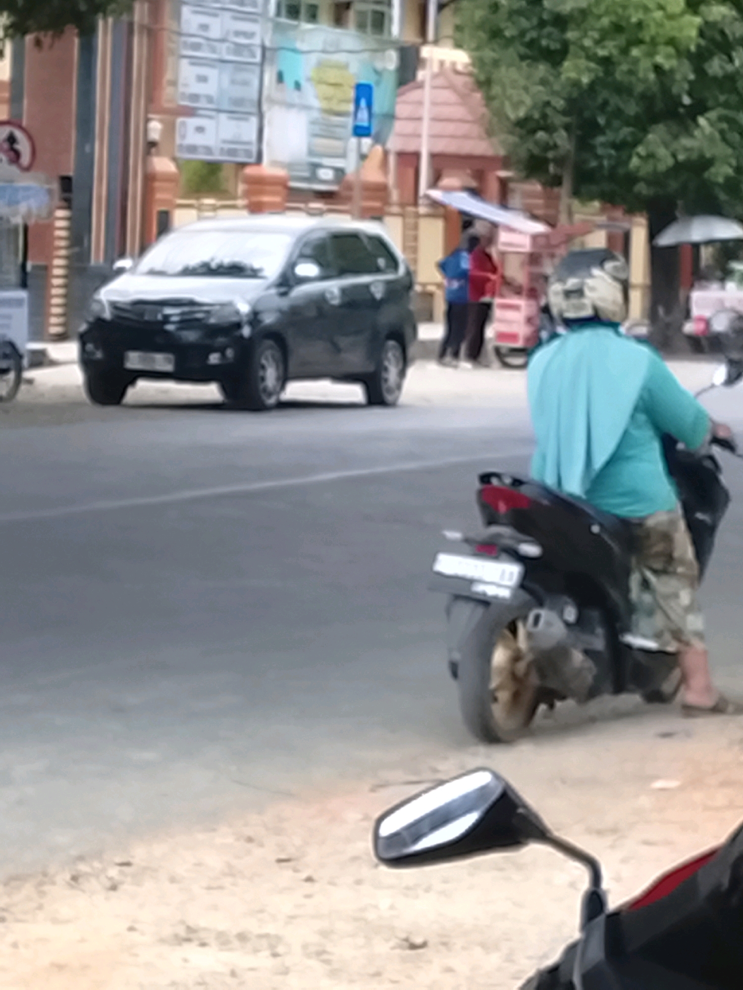 The image depicts a street scene in Slawi, Indonesia, with a focus on a motorcycle rider and a car. A person wearing a turquoise headscarf and camouflage pants is riding a dark-colored scooter. The scooter has a license plate that appears to be white with black text, and there is a visible exhaust pipe. In the background, a black car, possibly a minivan, is parked on the left side of the road. The car has a license plate that is partially visible. Further in the background, there are buildings, trees, and street signs, suggesting an urban environment. There are also other individuals and a food cart visible in the distance. The lighting suggests it is daytime.