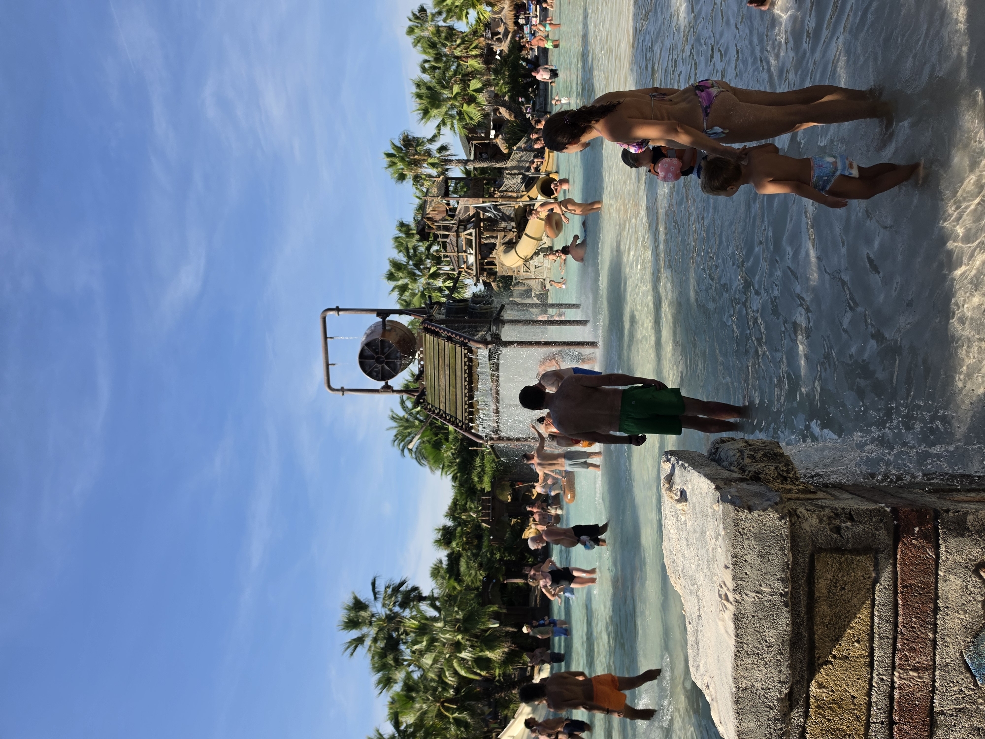 The image depicts a bustling water park scene on a sunny day, likely in Jesolo, Italy. Several people are enjoying the water, with some wading in the shallow pools and others appearing to be near a water slide structure. In the foreground, a woman in a bikini and a young child wearing swim diapers are standing in the water. The child has a flotation device around their waist. In the background, a wooden water slide complex with tubes is visible, surrounded by palm trees. More people are seen in and around the water, suggesting a lively atmosphere. The sky is a clear blue with some wispy white clouds, indicating fair weather. The sun is high in the sky, suggesting it is midday. The presence of palm trees and the overall ambiance strongly suggest a warm-weather resort or amusement park. No text is visible in the image.