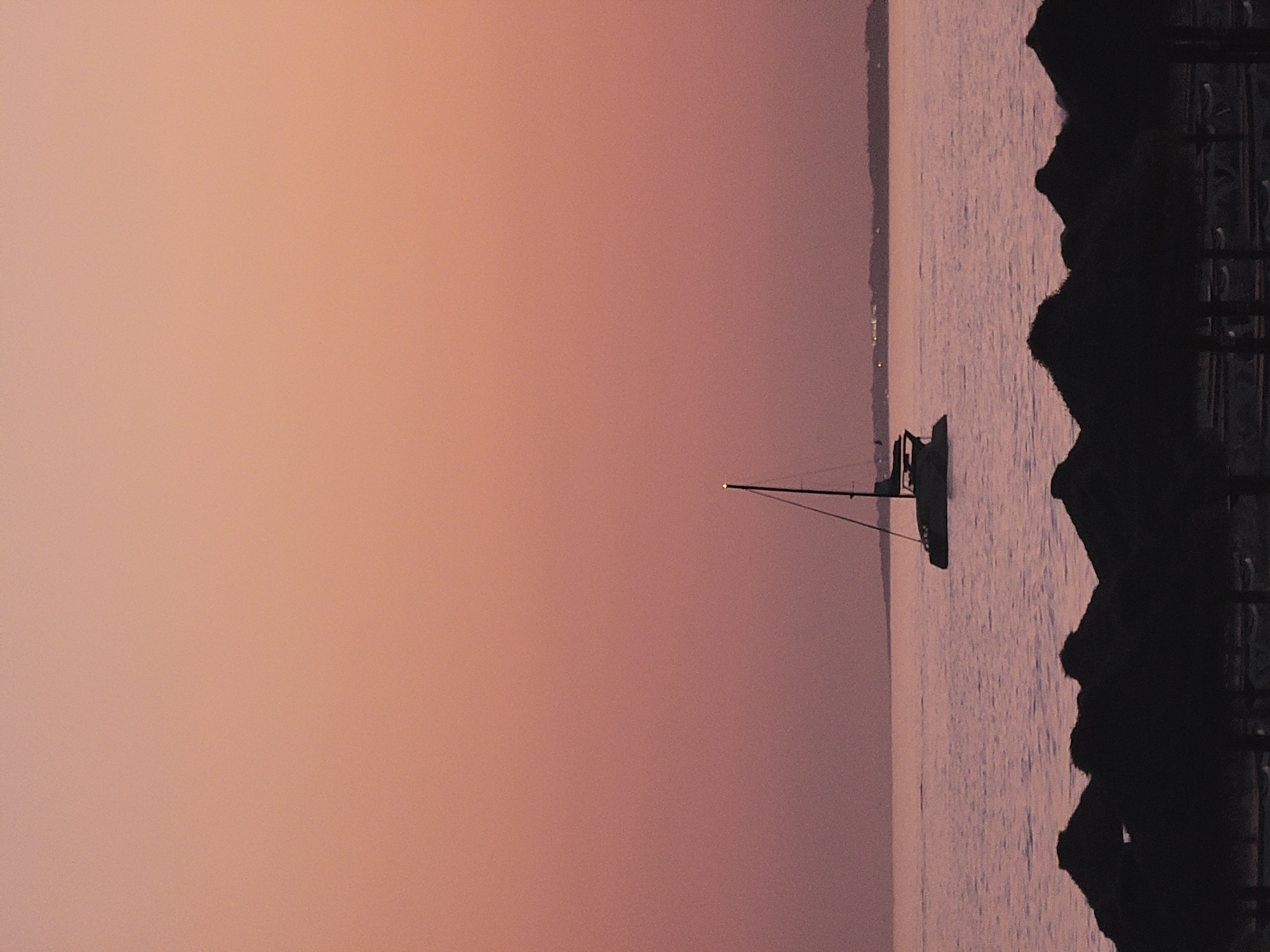 A catamaran sails on calm waters under a dusky sky. The catamaran, silhouetted against the horizon, features a tall mast with a single light at its apex. In the foreground, the dark silhouettes of thatched beach umbrellas create a distinct line across the bottom of the frame. The water reflects the soft hues of the sunset, which transitions from a pale pink at the top to a deeper rose towards the horizon. A distant landmass, possibly the Algarve coast near Quarteira, Portugal, is faintly visible. The overall scene is serene and evocative of twilight.