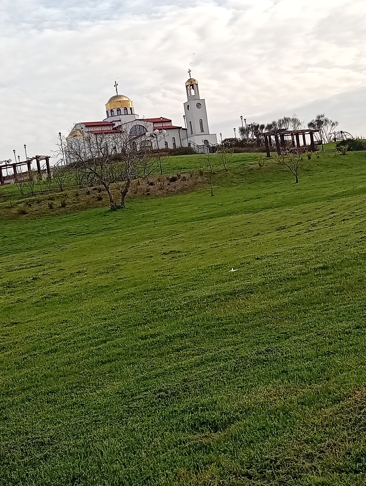 The image depicts a grassy hillside with a white and gold church complex at the top. The church features a golden dome and a separate, white bell tower. Several small trees and pergola-like structures are scattered along the slope. The sky is overcast with light clouds. The location is Unknown, Unknown.