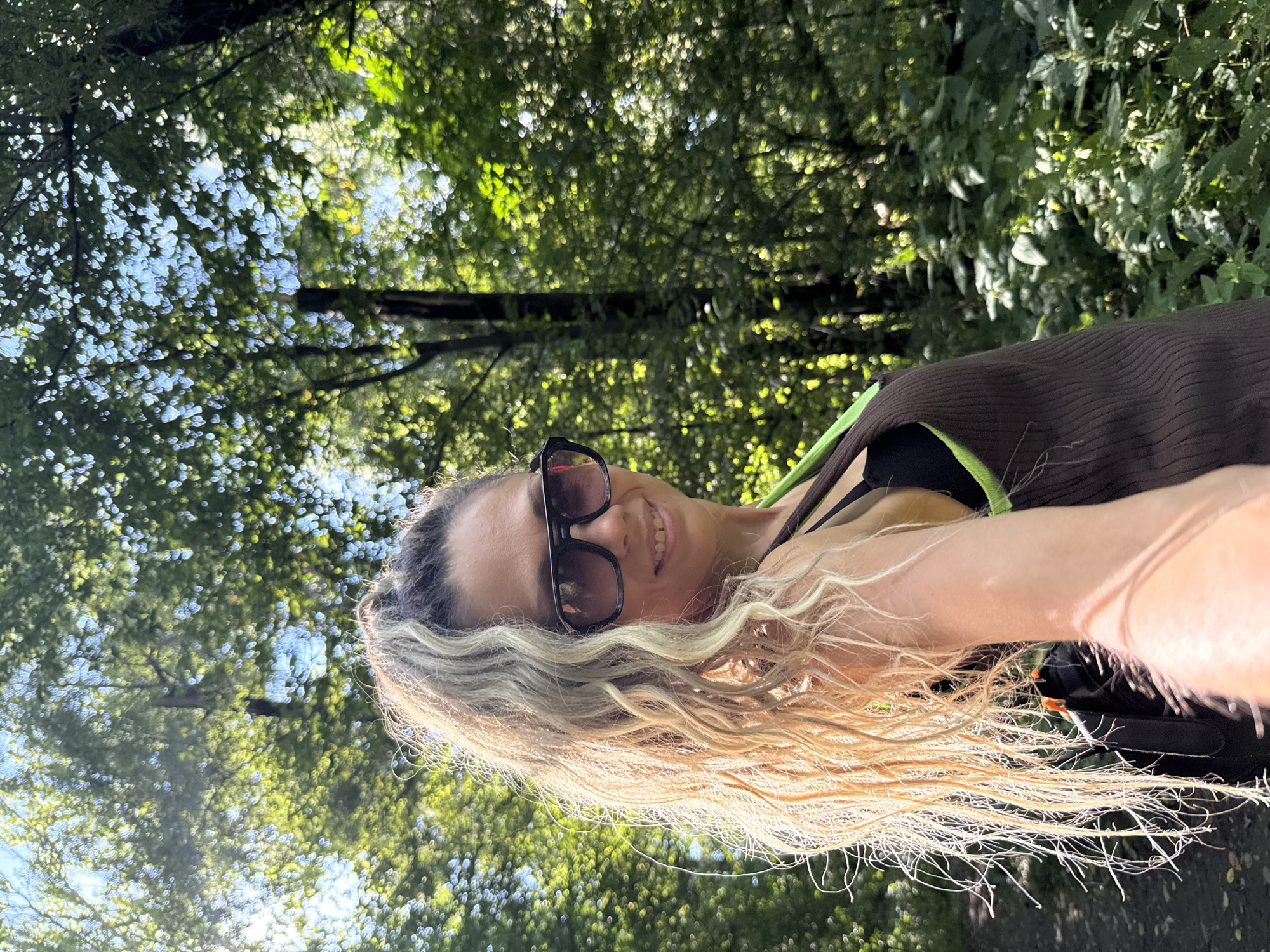 A woman is captured in a selfie taken outdoors in a wooded area with sunlight filtering through the trees. She has blonde hair pulled into a high ponytail and is wearing sunglasses and a brown sleeveless top over a black bra with neon green accents. The background consists of lush green foliage and tall trees, suggesting a natural, possibly park-like setting. The lighting indicates it is daytime.