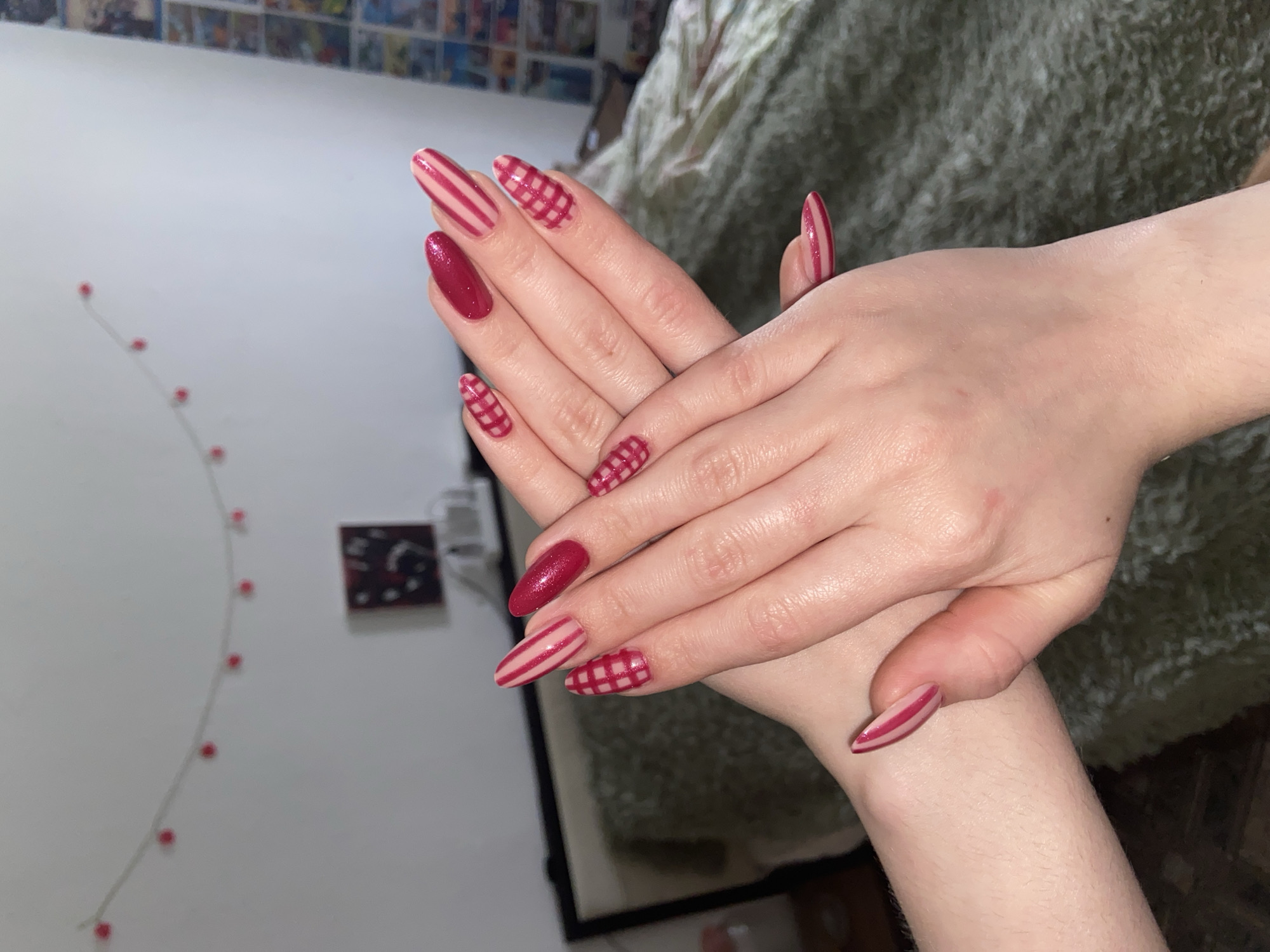 This indoor photograph, captured in Perm, Russia, focuses on a person's hands showcasing a detailed manicure. The hands, featuring fair skin, are gracefully posed one over the other, drawing attention to ten long, almond-shaped artificial nails. The nail art presents a mix of designs: some nails are painted a solid, glittery deep red, while others feature a light pink or nude base adorned with either distinct red stripes or a red and light pink/nude plaid/gingham pattern.
The background elements suggest a domestic setting. To the left, a string of small, round red lights hangs vertically against a clean white wall. Above this, a blurry shelf with various objects is visible. In the right half of the background, a textured, fuzzy greenish-grey blanket or fabric is draped, likely on a bed or sofa. A small, dark, out-of-focus object, possibly a framed picture, is also visible on the white wall. The lighting is consistent with an indoor environment. There is no visible text in the image. The primary activity depicted is the display of the elaborate nail art.