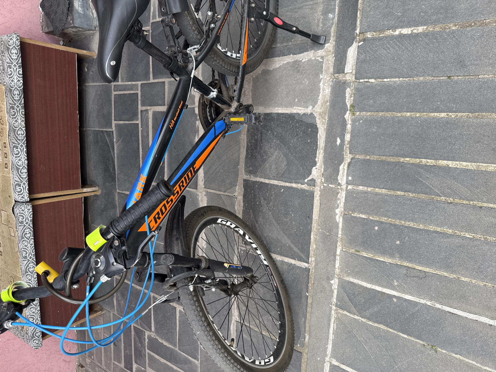 An eye-level, medium shot captures a black and orange mountain bicycle parked on a gray tiled sidewalk outdoors. The bicycle is positioned diagonally, with its front wheel closer to the viewer. The front wheel is black with silver spokes and a white stripe around the rim, with the word "GRAVITY" visible. The handlebars are black with yellow grips and blue brake cables. A yellow lock is attached to the handlebars. The frame of the bicycle is black with orange and blue accents. The word "CROSSRIDE" is emblazoned on the frame in orange letters with a blue outline. A black seat with a white circular logo is visible. The rear wheel is black with silver spokes and a black tire. A black kickstand is extended to the right of the rear wheel. The background features a wooden bench with a patterned fabric seat, and a pink wall behind it. The sidewalk is made of dark gray rectangular tiles with lighter gray grout lines.