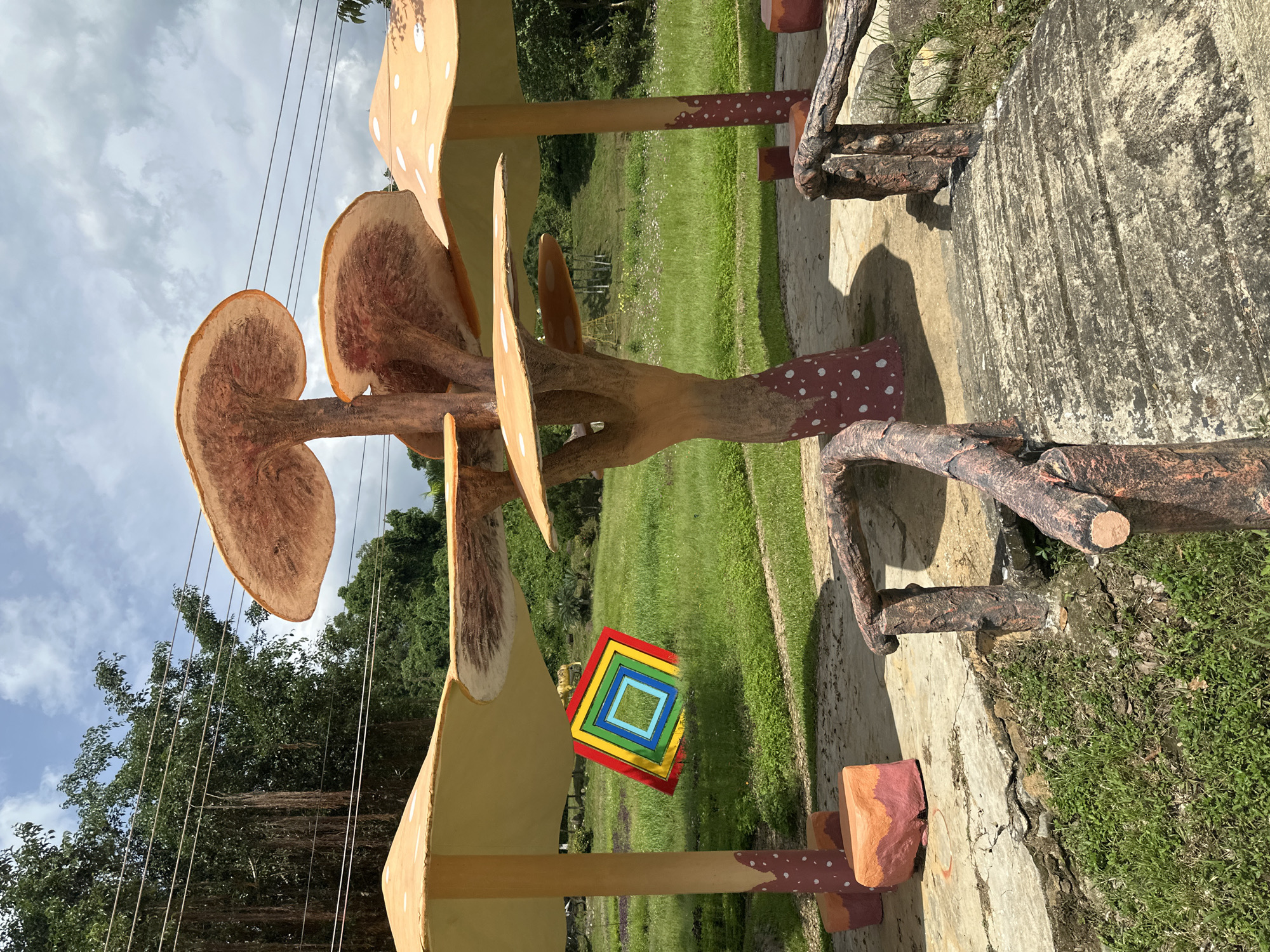 The image shows a decorative outdoor area with mushroom-themed sculptures. A central mushroom sculpture features a thick, textured trunk with multiple caps of varying sizes stacked atop each other, resembling a cluster. The caps are a light orange-brown color with darker undersides that show radiating lines. The base of the central mushroom and the stems of surrounding mushroom structures are decorated with red and white spots.
To the left and right of the central sculpture are two larger mushroom structures with wide, flat caps. The cap on the left is supported by a thick, spotted stem, and a colorful, square mosaic featuring concentric squares of red, yellow, green, blue, and white hangs from beneath it. The cap on the right is also supported by a spotted stem.
In the foreground, there is a small, rustic-looking bridge made of what appears to be faux wood material, with a textured surface. The bridge has railings made of the same material, designed to resemble tree branches. Two cube-shaped, multi-colored seating elements, red and orange, are placed on the ground near the base of the mushroom structures.
The environment is grassy and appears to be a park or garden. In the background, there are trees, a slight incline of land, and power lines running horizontally across the upper portion of the image. The sky is partly cloudy. The overall scene suggests a whimsical or fantasy-themed recreational space. The location is identified as Khánh Vĩnh, Vietnam.