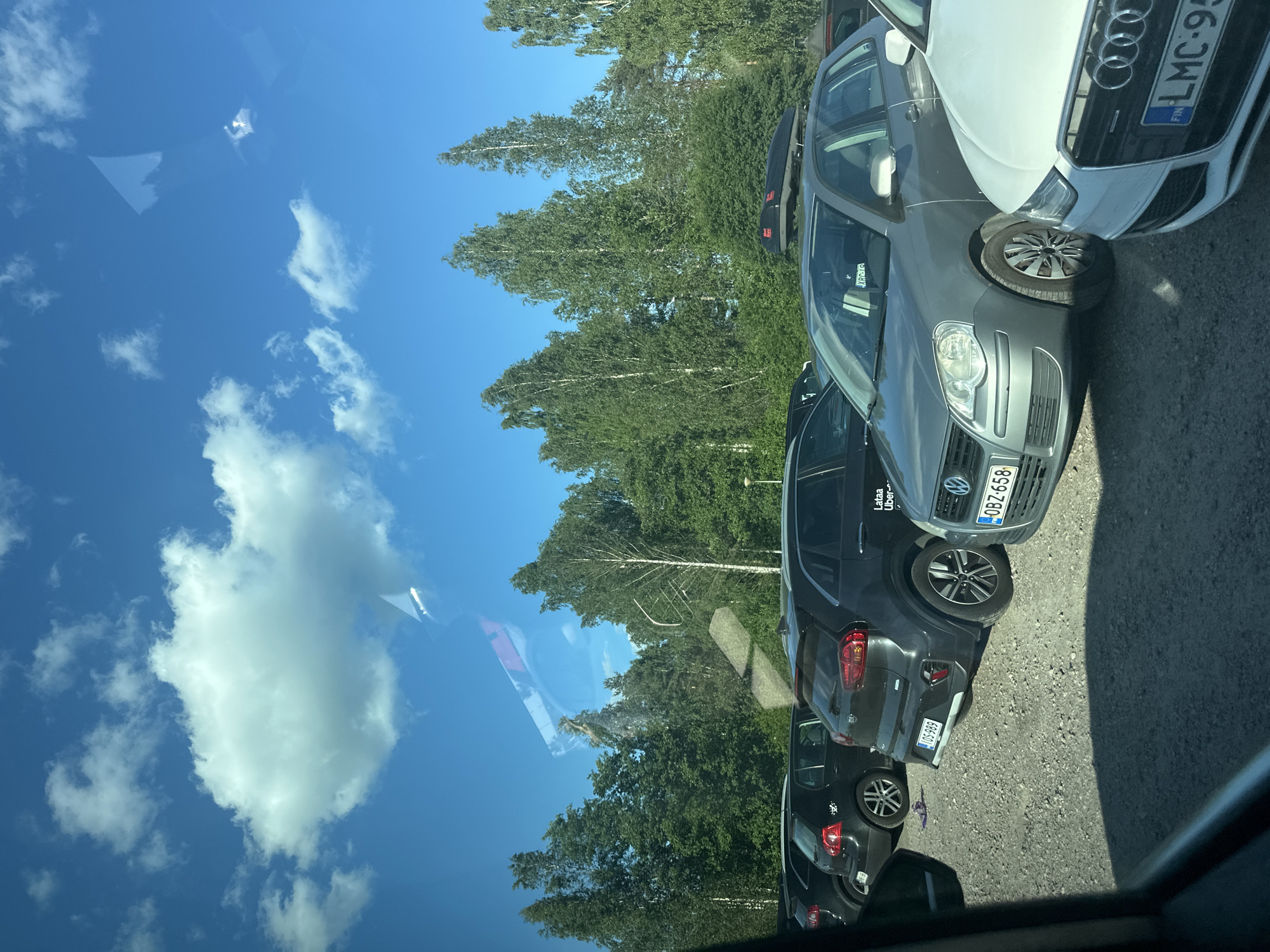 An outdoor, eye-level, full shot captures a parking lot filled with cars and surrounded by lush green trees under a bright blue sky with scattered white clouds. The cars, mostly dark-colored SUVs and sedans, are parked in a gravel lot. A few vehicles are visible in the foreground, including a white Audi with a Finnish license plate reading "LMC-95," a grey Volkswagen Polo with a license plate "OBZ-658," and a dark grey Kia SUV. The Kia's license plate is partially obscured but appears to read "IOS-989." Reflections of clouds and what appears to be car interior elements are visible on the windshields of some vehicles. The trees are tall and dense, creating a natural backdrop to the scene. The overall impression is a well-lit day in a suburban or rural setting.