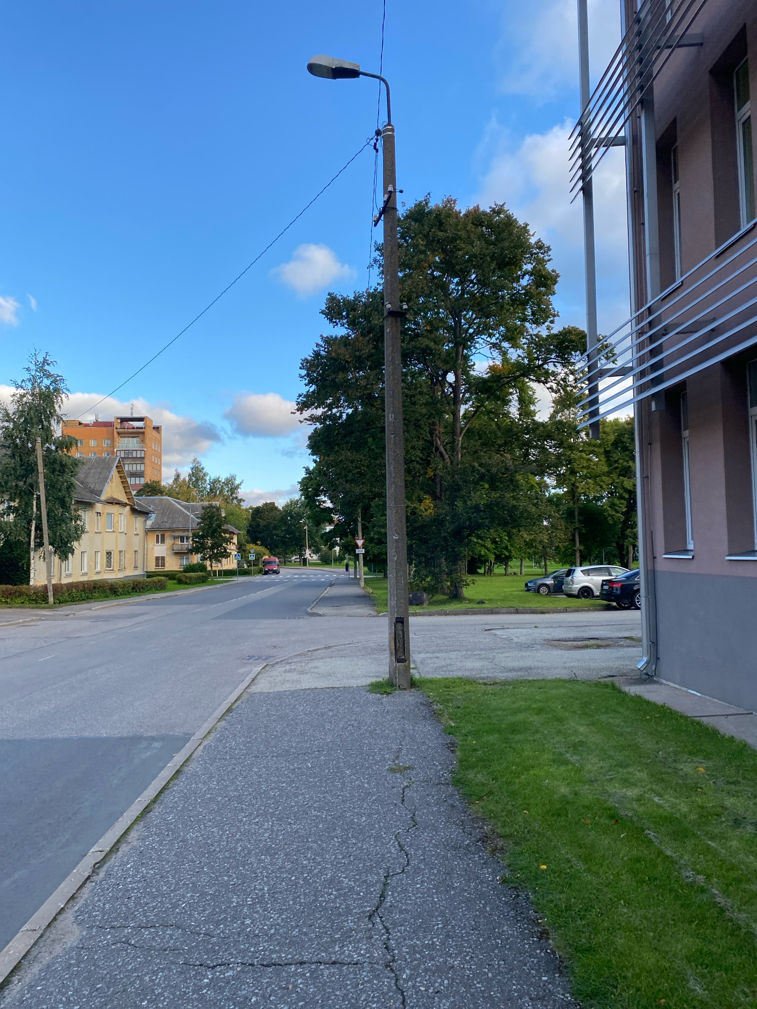 The image depicts a street scene in Kohtla-Järve linn, Estonia. In the foreground, a cracked asphalt sidewalk runs alongside a gray asphalt road. To the right of the sidewalk is a patch of green grass. A concrete utility pole with electrical wires extends upwards, topped with a street lamp. Adjacent to the grass is a modern building with a gray facade and horizontal metal accents on its upper exterior.
Across the road, on the left side, are residential buildings. The closest building is a two-story yellow house with a gray roof. Behind it, a taller, orange-brick apartment building is visible in the distance. Trees with green foliage line the street and are scattered throughout the background.
Several cars are parked on the grass area across the street from the modern building: a dark-colored sedan, a silver hatchback, and another dark-colored sedan. In the distance on the road, a red vehicle is traveling away from the camera. The sky is blue with scattered white clouds, suggesting daylight. The overall scene appears to be a residential or mixed-use area within the city.
