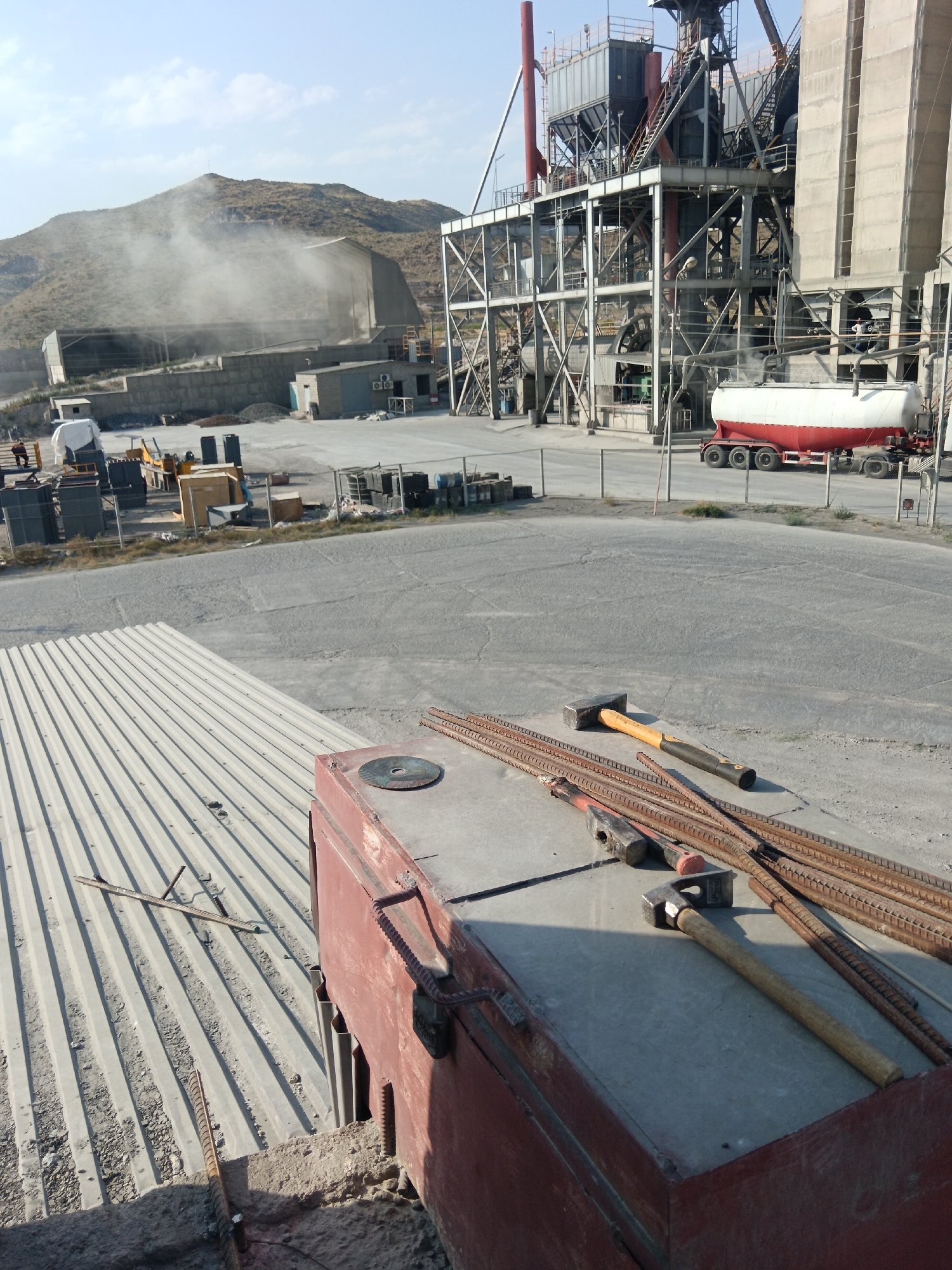 The image captures an industrial site, possibly a cement plant, located in Aragatsavan, Armenia. In the foreground, a red industrial container is visible, with rebar and hammers laid out on a concrete surface. A circular cutting disc is also present on the container. In the mid-ground, a large industrial facility with complex metal structures, silos, and conveyor belts dominates the scene. A white tanker truck with a red chassis is parked in front of the facility. The background features a dusty, mountainous landscape, with a large building partially obscured by dust or smoke. Several smaller structures, fences, and various construction materials like concrete blocks and rebar are scattered throughout the site. The sky is clear with some light clouds, indicating daytime. No people are clearly identifiable in the image.