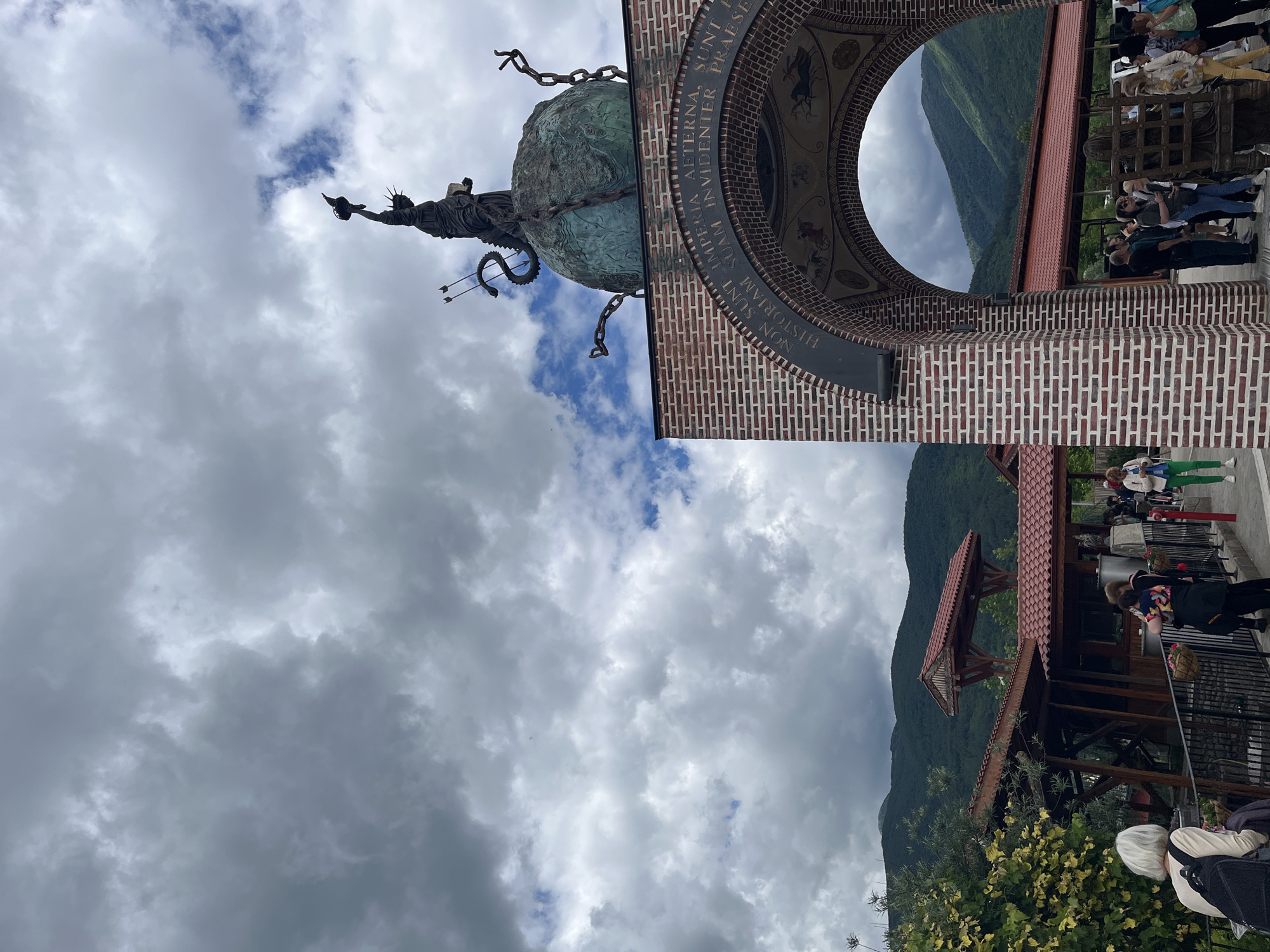 This is a medium shot, outdoor, eye-level photograph taken on a cloudy day. The scene depicts the entrance to a memorial complex in Skobelevo, Bulgaria. In the foreground, a brick archway frames a view of the memorial and surrounding landscape. Above the arch, a statue of a figure holding a torch stands atop a globe, which is encircled by chains and a serpentine creature. The globe appears to be made of a verdigris-patinated metal. The archway itself is adorned with text in Latin, which reads "NON SUNT IMPERIA AETERNA, SUNT HISTORIAM SUAM. INVIDENTER PRAESE". Within the arch, a fresco depicts chariots pulled by horses.
Beyond the archway, several people are visible. Some are standing in a queue, looking towards the memorial. Others are walking through the complex, and a few are gathered in small groups. Among the visible individuals are a woman with gray hair wearing a beige top and a black backpack, a woman holding a child, and people dressed in casual attire, including green pants and white tops.
The background reveals a scenic mountainous landscape covered in lush green vegetation. Several wooden structures with red-tiled roofs are also present, suggesting a park or historical site. The sky is filled with dramatic, dark, and gray clouds, indicating overcast weather.
