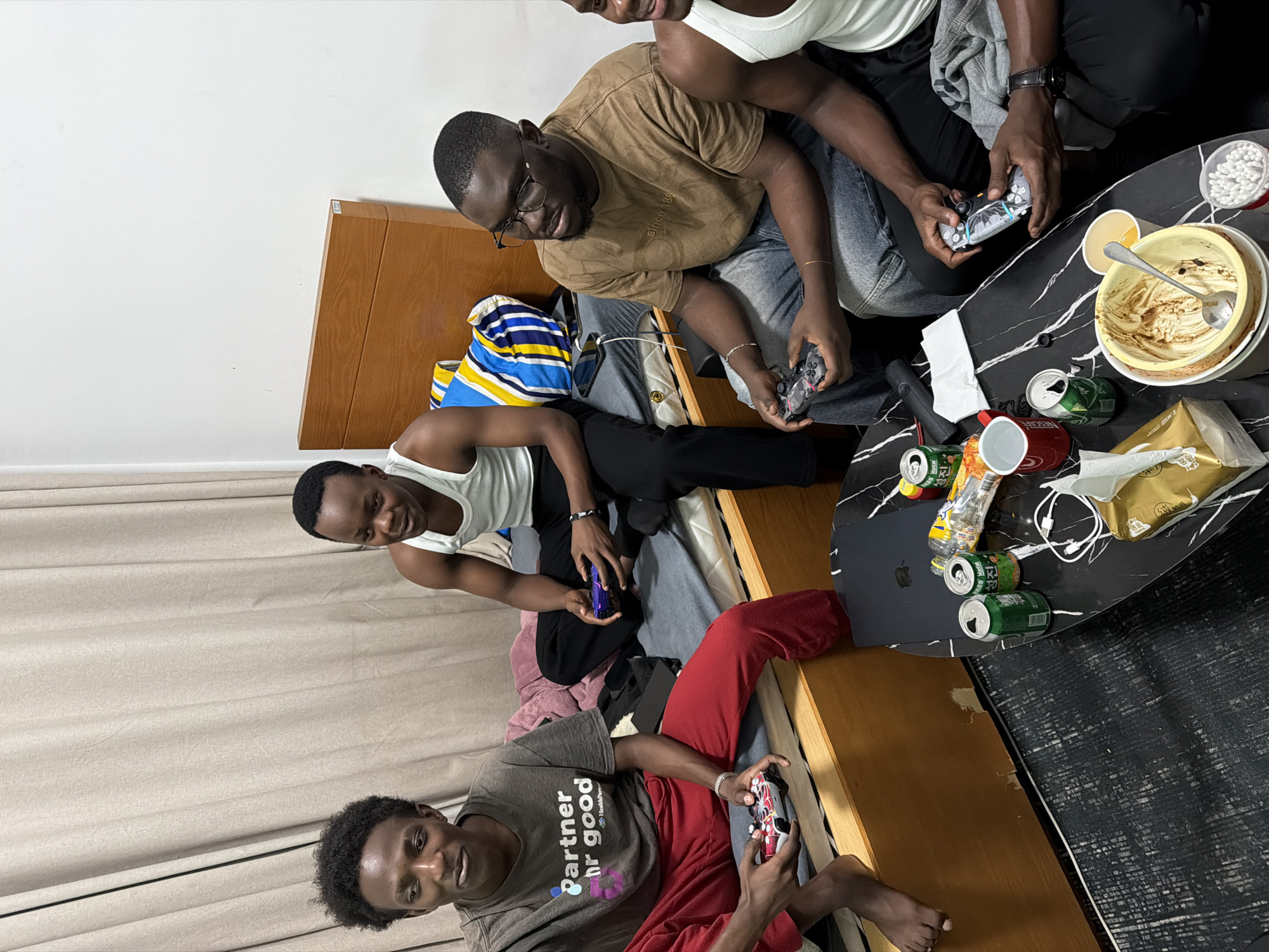 Four young Black men are gathered in an indoor room, engaged in playing video games. The scene is shot from a slightly elevated, rotated perspective, showing the men in various seated positions around a low black table with a white marble pattern.
Two men are seated on a bed, one in a white tank top and black pants, holding a video game controller, and another in a brown t-shirt and glasses, also holding a controller and looking down at it. A third man, wearing a white tank top, sits towards the bottom left of the bed, holding a controller and looking towards the camera with a slight smile. The fourth man is seated on the floor to the left, wearing a grey t-shirt with a "Partner hr good" logo (featuring a heart graphic and "HealthP") and red pants, holding a patterned controller and smiling at the camera. Another man's brown t-shirt has small text reading "Since 1986".
The black table is cluttered with various items, indicating a casual gaming session with snacks and drinks. There are two empty or partially empty food bowls, one yellow and one white, with a spoon in one, suggesting they have just eaten. Numerous empty or crumpled beverage cans (green, red, silver, and a transparent bottle) are scattered across the table. Other items include a container of cotton buds, white wired earphones, and a few pieces of discarded paper or packaging.
The room features a simple decor with a wooden bed frame or headboard, light-colored curtains in the background, and a grey patterned rug or carpet on the floor. The overall atmosphere appears relaxed and social, typical of friends spending leisure time together. The location context indicates Jiangning District, China.