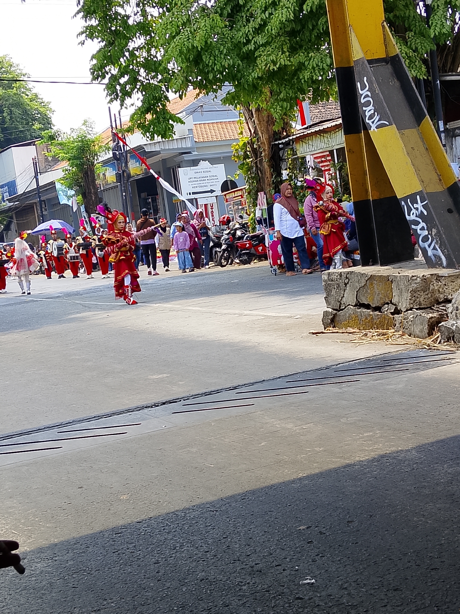 In Kertosono, Indonesia, a street parade is in progress. A group of children in red and gold uniforms with elaborate headdresses march in formation, some carrying drums. A young girl in the foreground, also in a red and gold costume, holds a baton. Spectators in casual clothing line the street, some watching the parade, others engaged in conversation. A large yellow and black striped pole, possibly a traffic signal or barrier, stands prominently on the right side of the frame. Buildings and trees line the background, indicating an urban setting. The scene captures a vibrant community event on a sunny day.