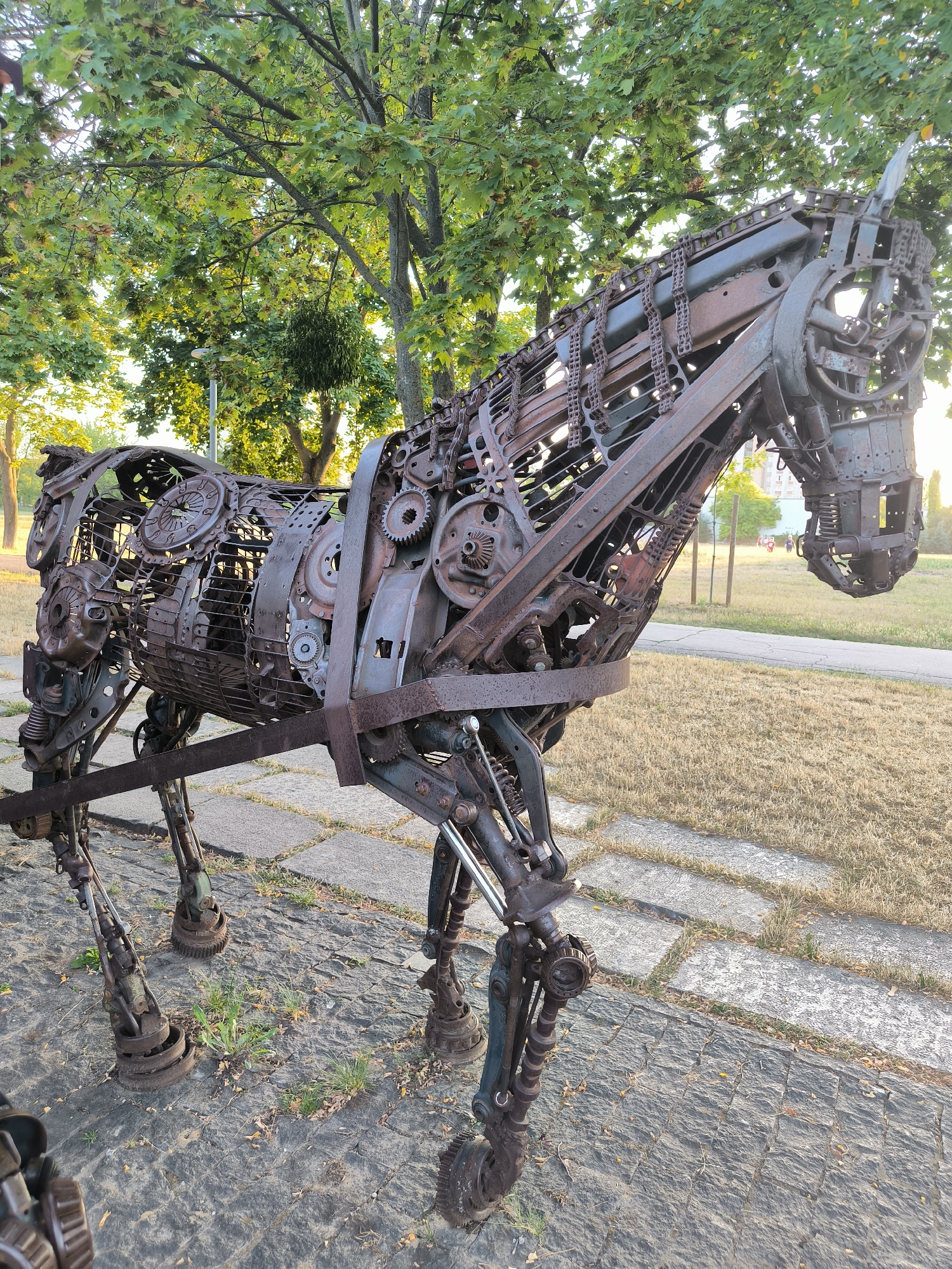 A large, rustic metal sculpture of a horse stands in a park. The horse is constructed from various scrap metal parts, including gears, chains, and automotive components, giving it a steampunk aesthetic. It is positioned on a paved pathway made of stone slabs, with dry grass and trees in the background. The lighting suggests it is late afternoon or early evening, with the sun casting a warm glow. The sculpture is the central focus, with no people actively interacting with it in the frame. The environment appears to be a public outdoor space in Chișinău, Moldova, likely a park or recreational area.