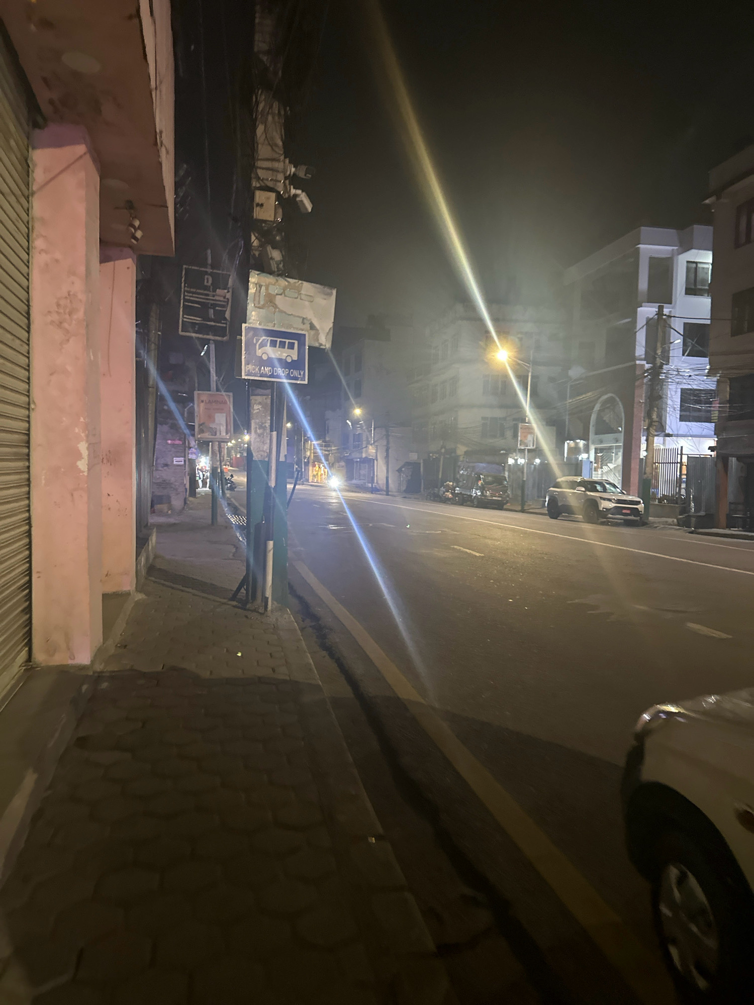 This image depicts a street scene at night in Kathmandu Metropolitan City, Nepal. The perspective is from a sidewalk facing a street. On the left, a light pink building with a metal roll-up door is visible, with a thick, light pink pillar in the foreground. A sidewalk paved with hexagonal stones runs alongside the building. A lamppost with several signs, including a bus stop sign indicating "PICK AND DROP ONLY," is positioned on the sidewalk.
Across the street, a paved road with yellow lane markings is visible. Several vehicles are parked or driving on the road. A white SUV is parked on the right side of the road, and a darker vehicle, possibly a van, is parked further down on the left. Buildings of varying heights and architectural styles line the opposite side of the street, illuminated by streetlights and internal lighting. The overall atmosphere is dimly lit, with a slight haze or fog present in the air. Electrical wires are prominently strung overhead.