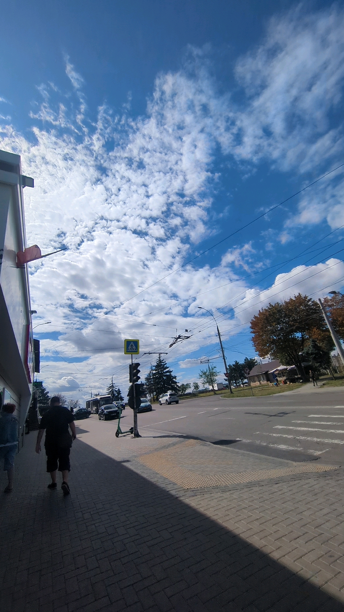 The image depicts a street scene on a sunny day with scattered clouds. The perspective is from a sidewalk looking towards a road and intersection. In the foreground, on the left, is the side of a building with a partial view of its facade. Two individuals are walking away from the camera on the sidewalk. One person, wearing a black t-shirt and dark shorts, is closer to the camera. Another person, partially visible on the far left, wears a denim jacket and light-colored shorts.
The sidewalk is paved with interlocking bricks, and a section of the pavement is marked with tactile paving. Further down the sidewalk, a traffic light and a pedestrian crossing sign are visible, indicating an intersection. A green electric scooter is parked near the traffic light pole.
The road features a crosswalk and several vehicles, including a trolleybus, a dark sedan, and a white car further down the road. The intersection appears to be a typical urban junction with overhead power lines for the trolleybus system.
In the background, there are trees, some with autumn foliage, and buildings. The sky is a bright blue with fluffy white clouds. The scene suggests an active urban environment with pedestrian and vehicular traffic. The presence of a trolleybus and the architectural style of the building hint at a location in Eastern Europe, potentially Chișinău, Moldova, given the context provided.