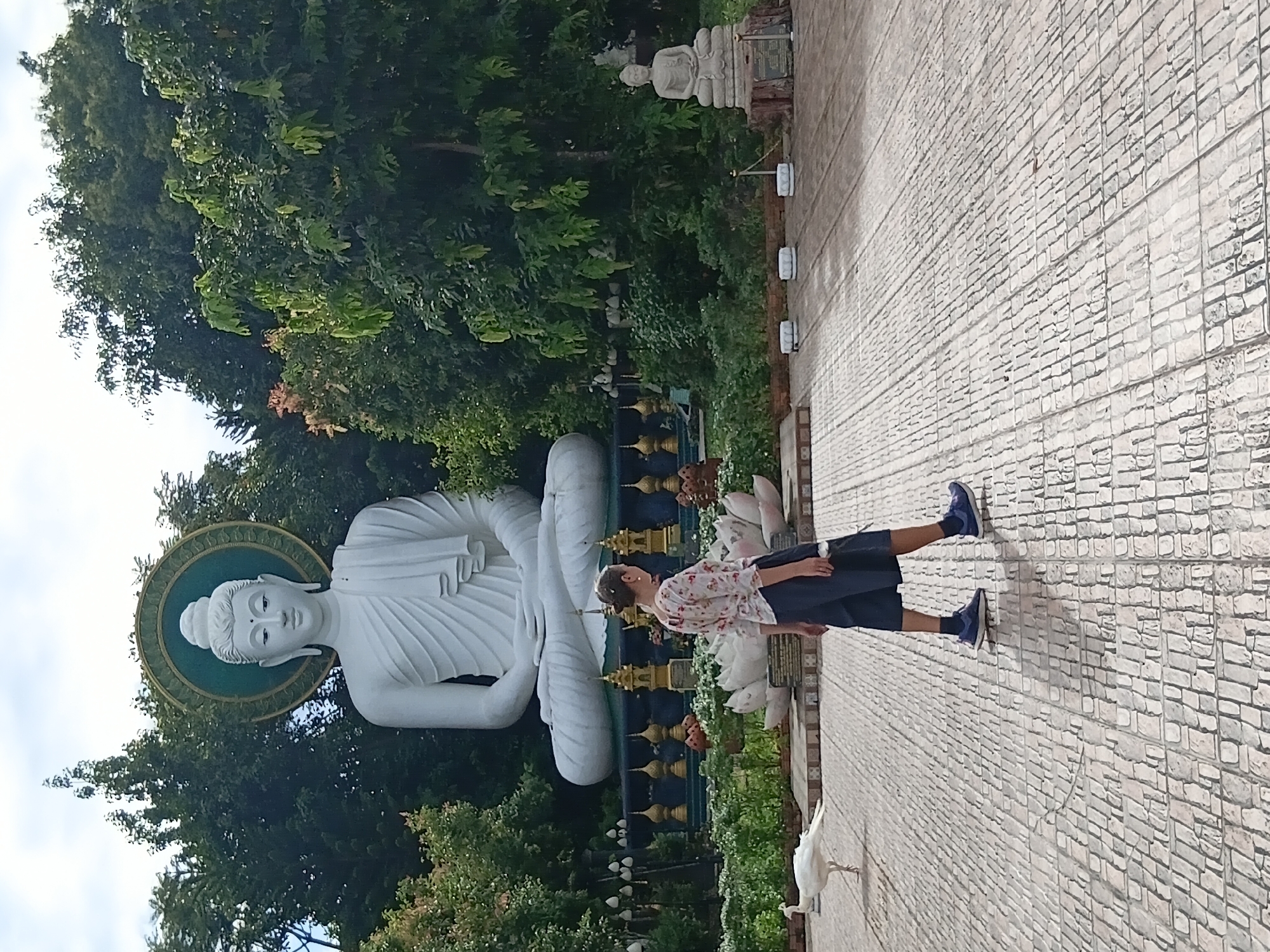 The image depicts a paved courtyard with a large, white Buddha statue seated in the background, surrounded by lush green trees. To the left of the Buddha, a white peacock is walking on the paved surface. A person, appearing to be female, is walking from left to right in the foreground, casting a shadow on the ground. The person is wearing a patterned shirt and dark shorts. The environment suggests a religious or cultural site, possibly a temple or sanctuary, consistent with locations like Ban Ruammit, Thailand, known for its natural beauty and spiritual significance. The lighting suggests it is daytime, with the sun casting distinct shadows. The overall scene conveys a sense of tranquility and cultural ambiance.