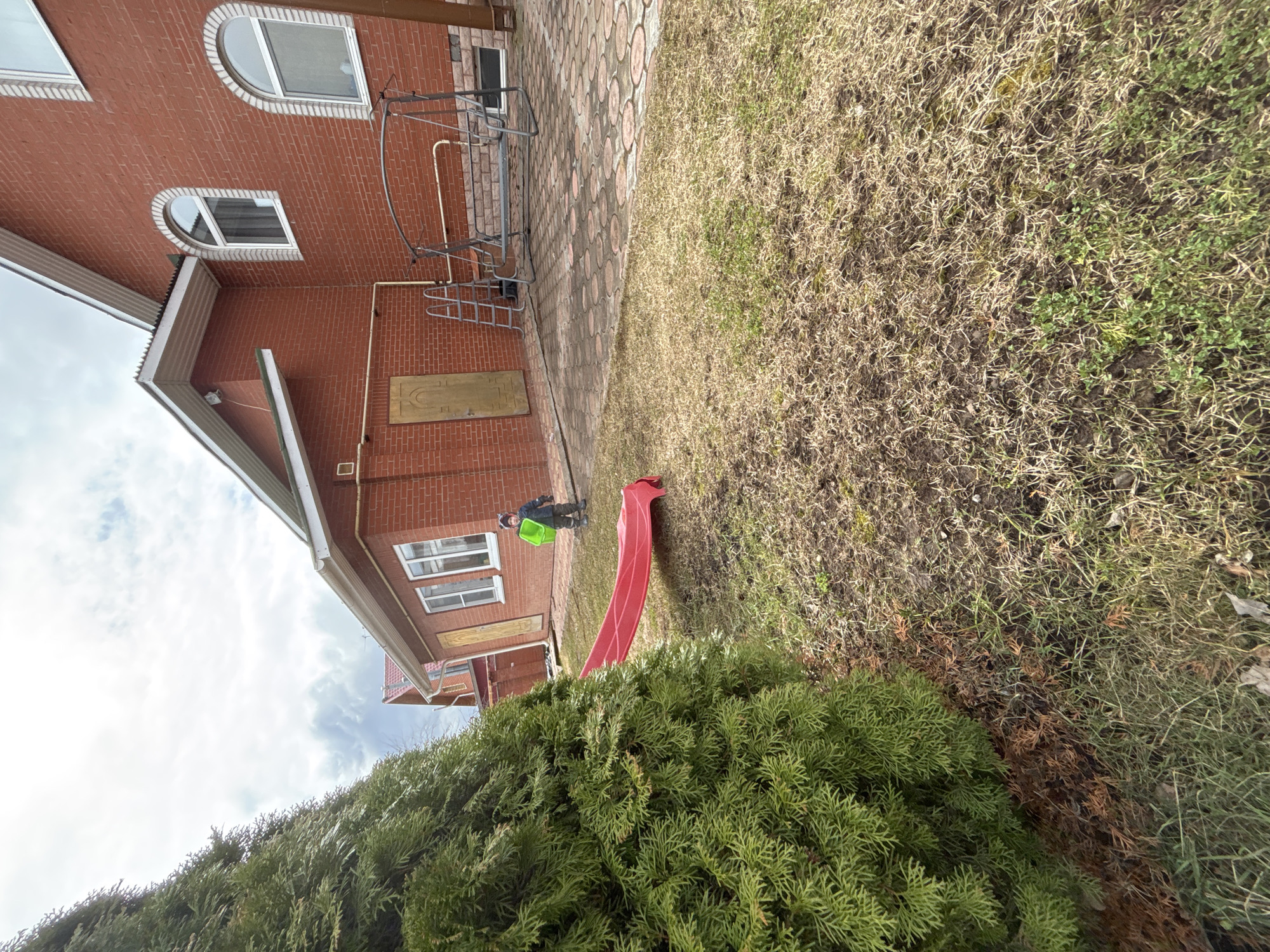 A sideways shot depicts a red brick building and a child with a green bucket in Ферма, Russia. The building has white-framed windows, including arched ones. A metal staircase is affixed to the side of the building. A red slide rests on the lawn, partially obscured by a dark green bush in the foreground. A young child wearing a dark jacket is near the slide, holding a green bucket. The lawn is a mixture of brown and green grass. The sky is cloudy. The location is Ферма, Russia.