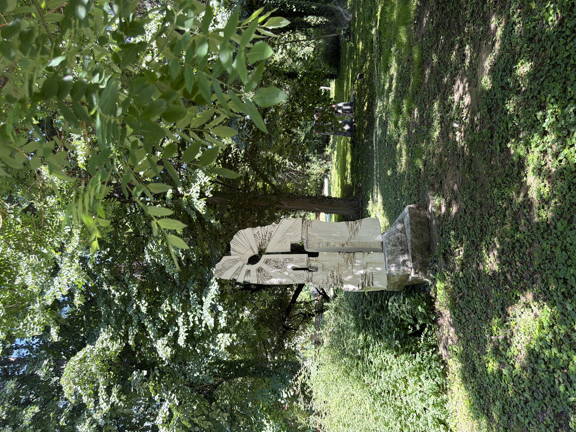 The image captures a modern abstract sculpture made of white stone, positioned in a park-like setting. The sculpture, with its angular and fragmented design, is situated on a stone base, surrounded by lush green foliage. In the background, dense trees create a shaded canopy, and a grassy area is visible, dotted with sunlight and shadows. Further in the background, a small group of people can be vaguely seen sitting or standing, partially obscured by the trees. The overall environment suggests a public outdoor space. Based on available information, this sculpture is located in the City Park of Pazardzhik, Bulgaria, and is known as "The Eternal Flight" by the sculptor Georgi Chapkunov.