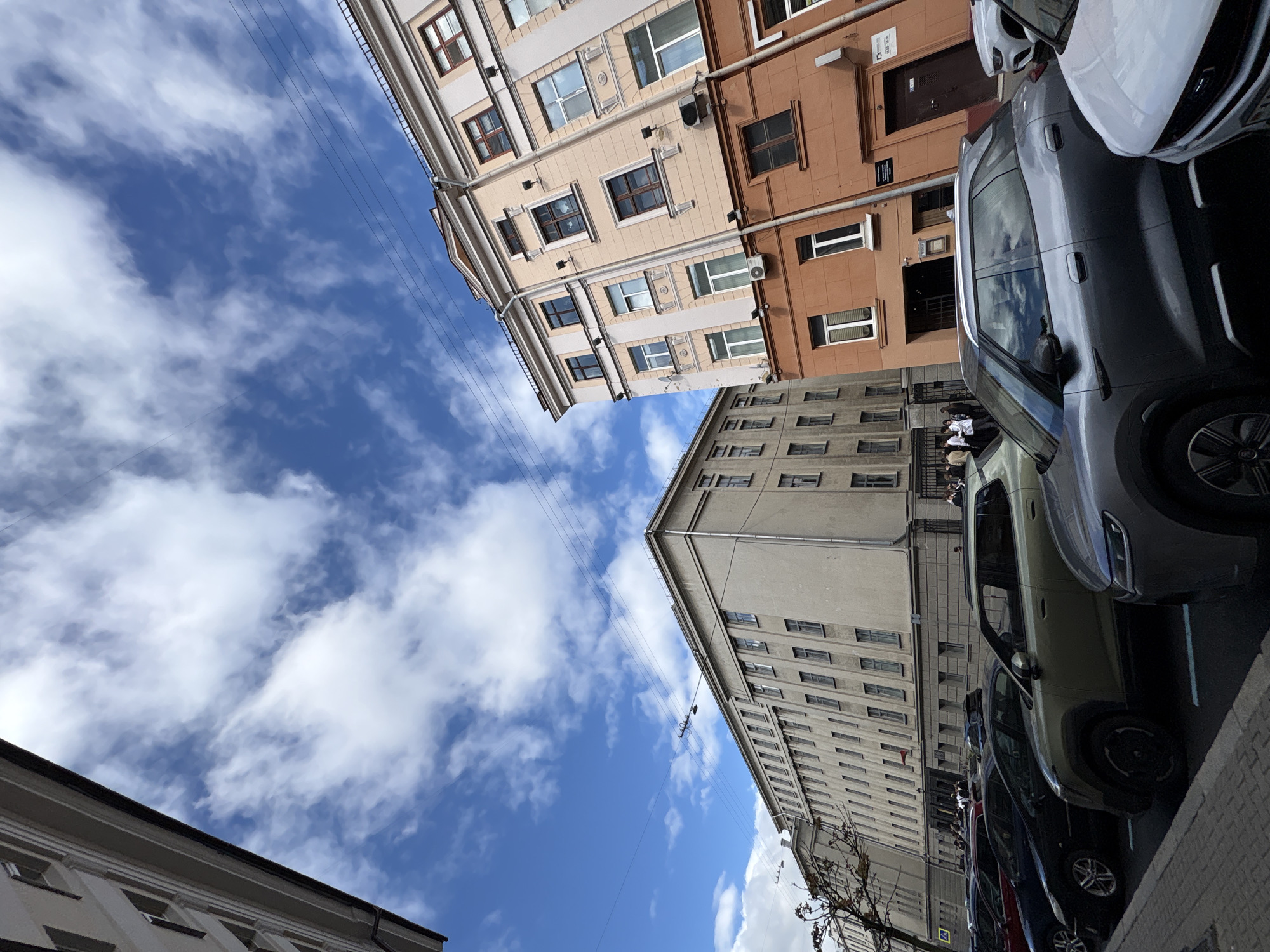 The image, taken at an unusual angle, shows an urban street scene in Minsk, Belarus. The view is angled upwards towards the sky filled with scattered white clouds. Two buildings are visible on either side of the frame. The building on the left is light beige and brown with multiple rectangular windows. The building on the right is grey. A row of parked cars is seen on the right side of the frame. The sidewalk runs along the base of the buildings. A few people are visible, seemingly standing near a building. Power lines run across the open sky space. Bare tree branches are also visible at the bottom right.