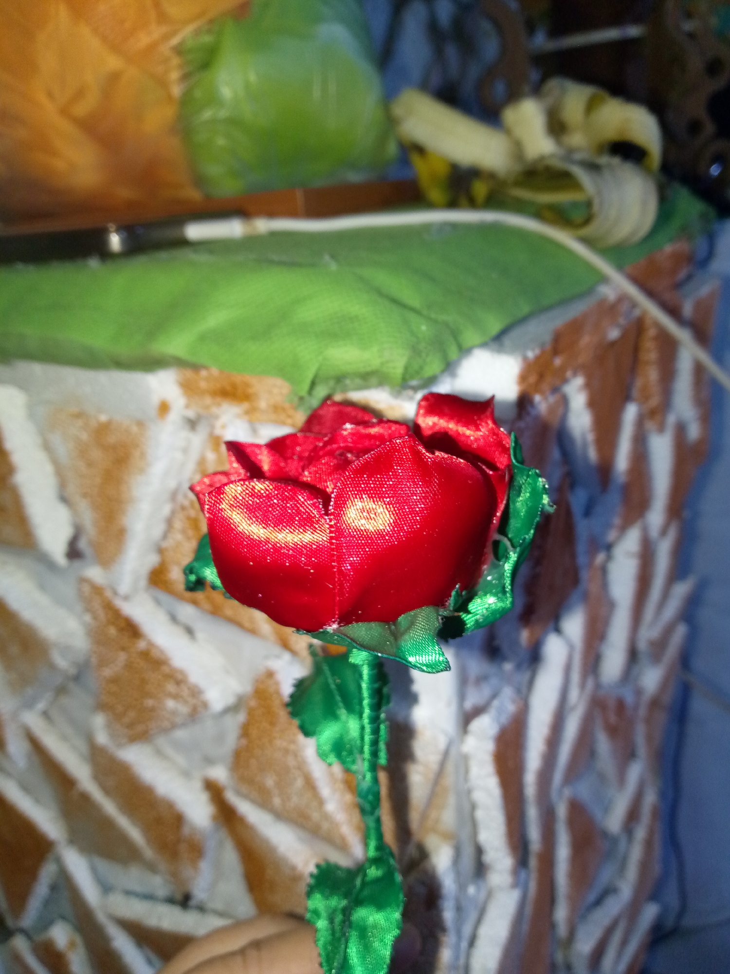 A close-up photograph depicts a red artificial rose with green fabric leaves and stem. The background is blurred, showing a wall with a mosaic pattern of triangular tiles in brown and white. Behind the wall, there is a blurry green cloth and discarded banana peels, along with a partially visible clear plastic bag and an orange package. The rose is positioned in the foreground, seemingly held by a hand slightly visible at the bottom left of the frame.
There is no verifiable evidence in the photo to suggest that this image was taken in Juan Griego, Venezuela.