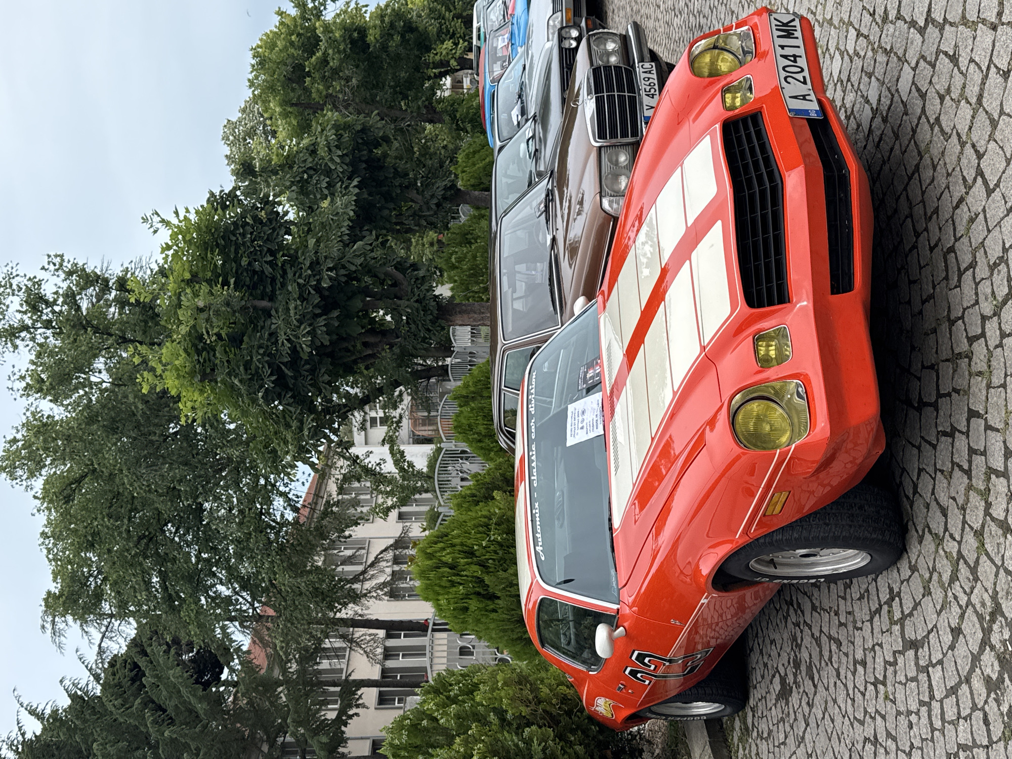 In Tsarevo, Bulgaria, a line of classic cars is parked on a cobblestone street. The foreground features a bright orange Chevrolet Camaro with white racing stripes and the number "22" on the driver's side door. Yellow headlights and a black grille are visible. Behind it, a brown Mercedes-Benz sedan is parked, displaying a license plate that reads "V 4569 AC". Further back, other classic cars of various colors are lined up. Lush green trees and a building with white siding and red-tiled roofs form the backdrop. The sky is overcast.