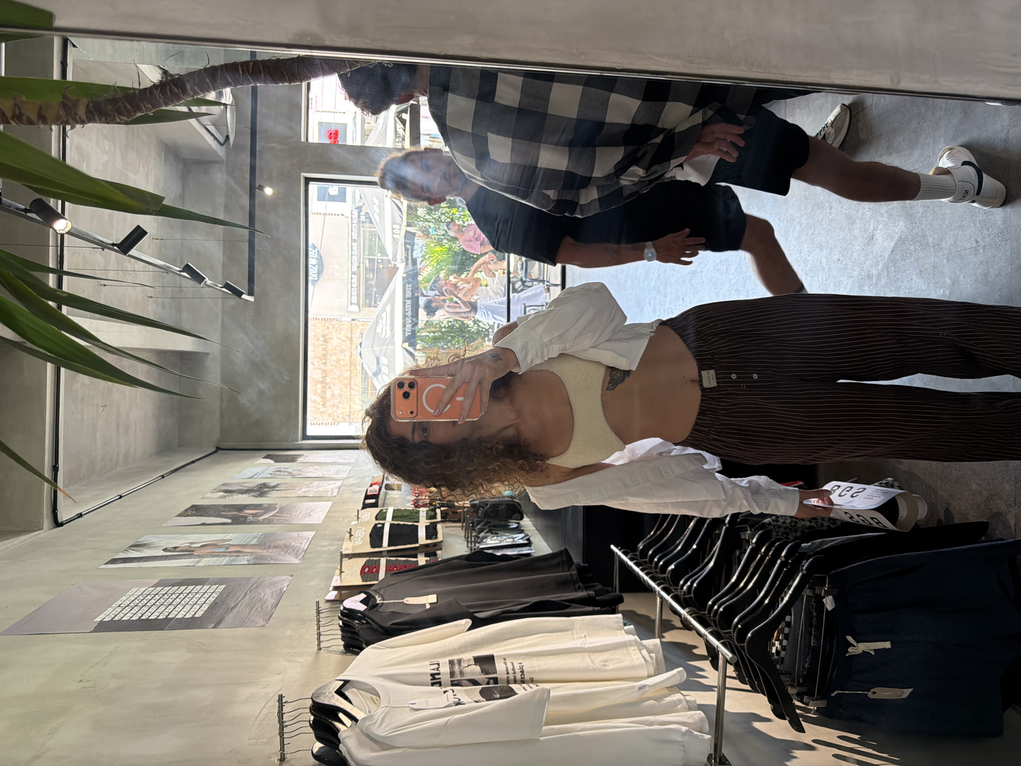 A person is taking a mirror selfie inside a clothing store in North Kuta, Indonesia. The selfie includes a man reflected in the mirror standing near the person. The man is wearing a plaid shirt and dark shorts. The store's interior has a concrete floor with a row of large photographs or posters on the floor. Clothes are displayed on racks, and a potted plant is visible near the ceiling. Outside the store, buildings and pedestrians are visible through the store's large windows.
