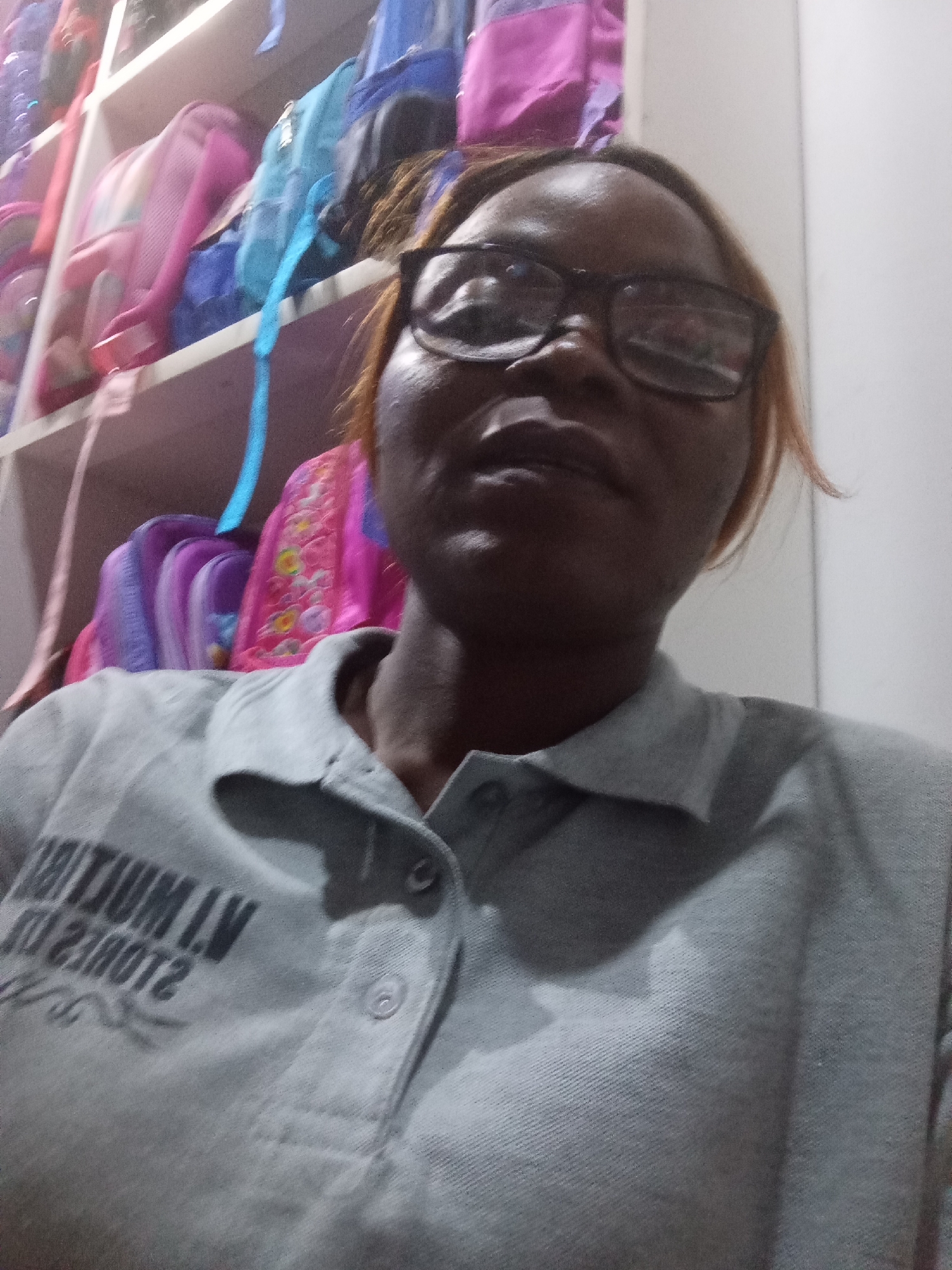 The image shows a woman wearing glasses and a grey polo shirt with lettering that appears to be "IVIUM I.V 21DUOTZ". She is looking at the camera. In the background, shelves are filled with colorful backpacks. The scene is indoors and appears to be a retail or storage space for school supplies. The location in Howayi, Nigeria is not discernible from the image itself.