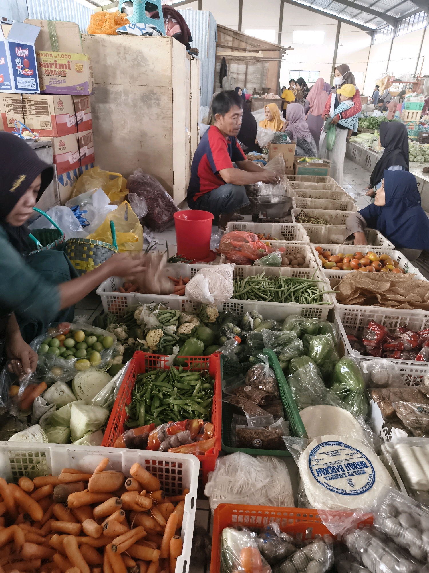 This image captures a bustling outdoor market scene in Sukorejo, Indonesia, likely a traditional wet market given the abundance of fresh produce. In the foreground, a woman wearing a black hijab and green clothing is seen tending to her stall. She is interacting with a bag of produce, possibly weighing or packaging it. Adjacent to her, a man in a red and blue shirt is also engaged with items on his stall, which appears to be set up with white plastic crates filled with various vegetables and fruits.
The market floor is tiled. Behind the stalls, more people are visible, some wearing hijabs and traditional clothing, suggesting a predominantly Muslim community. The market stalls are laden with a diverse array of fresh produce, including carrots, green beans, tomatoes, cabbages, cauliflower, and other leafy greens, all neatly arranged in plastic crates. Some items are bagged, while others are displayed loose. In the background, there are structures that appear to be covered market stalls, with a corrugated metal roof visible. The overall atmosphere suggests a busy commercial activity with people buying and selling goods. The presence of packaged food items on shelves in the background also indicates a wider range of products available.